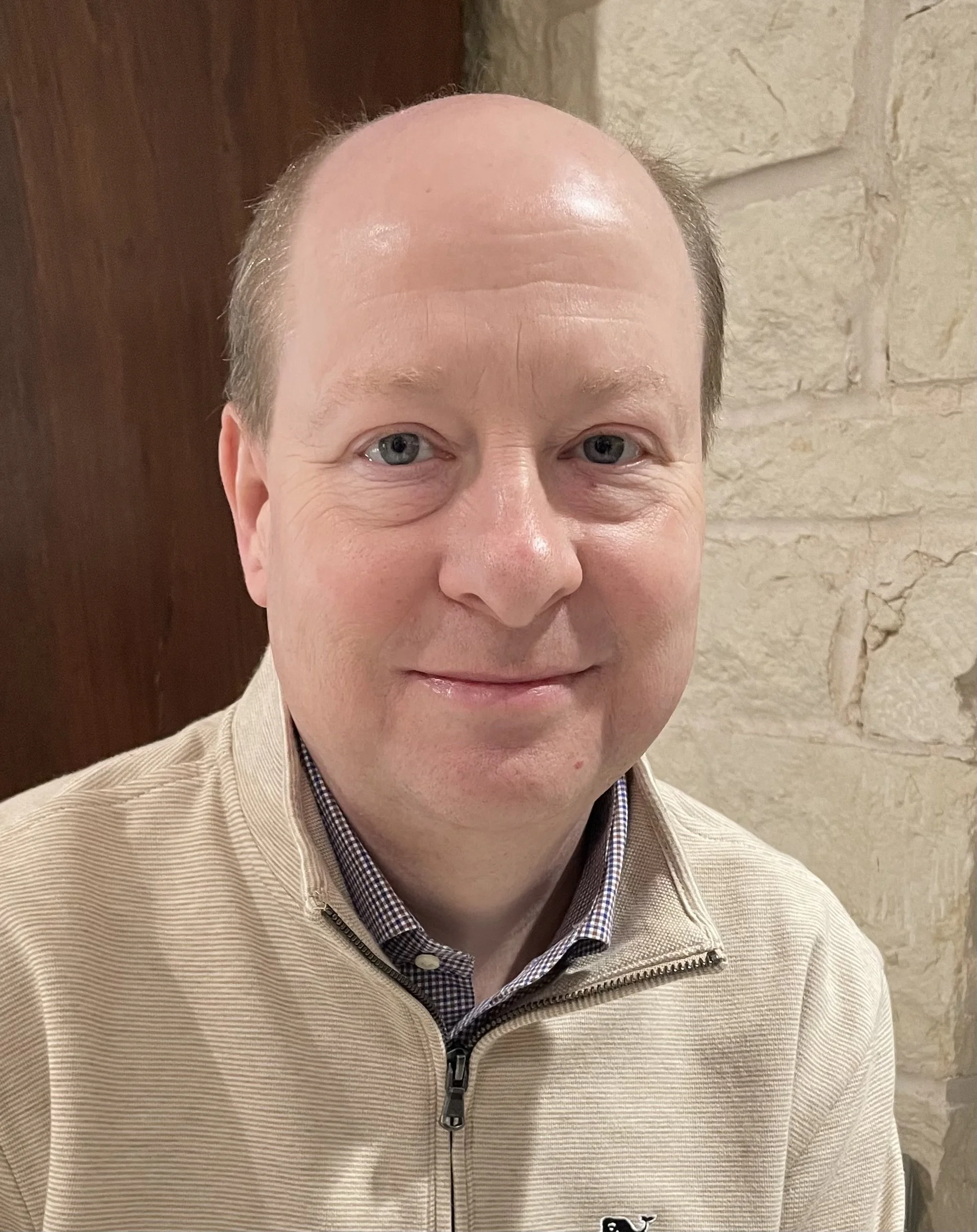 A man with light skin, short light brown hair, and blue eyes smiling, wearing a beige zip-up jacket over a checkered shirt, posed in front of a stone wall and wooden door.
