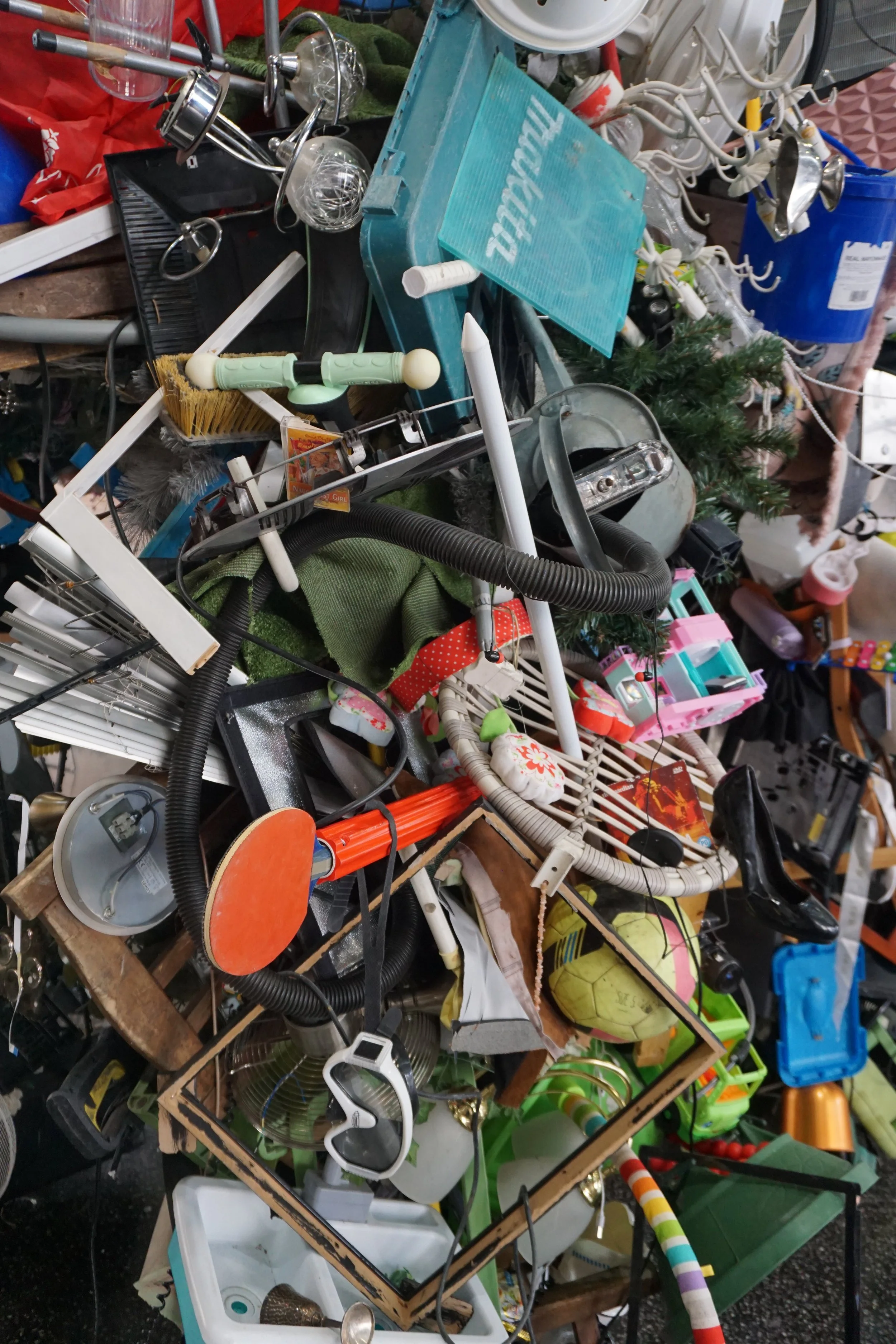 A cluttered assortment of miscellaneous household and decorative items including lighting fixtures, a small artificial Christmas tree, kitchen utensils, toys, and various other objects arranged on a multilevel display.