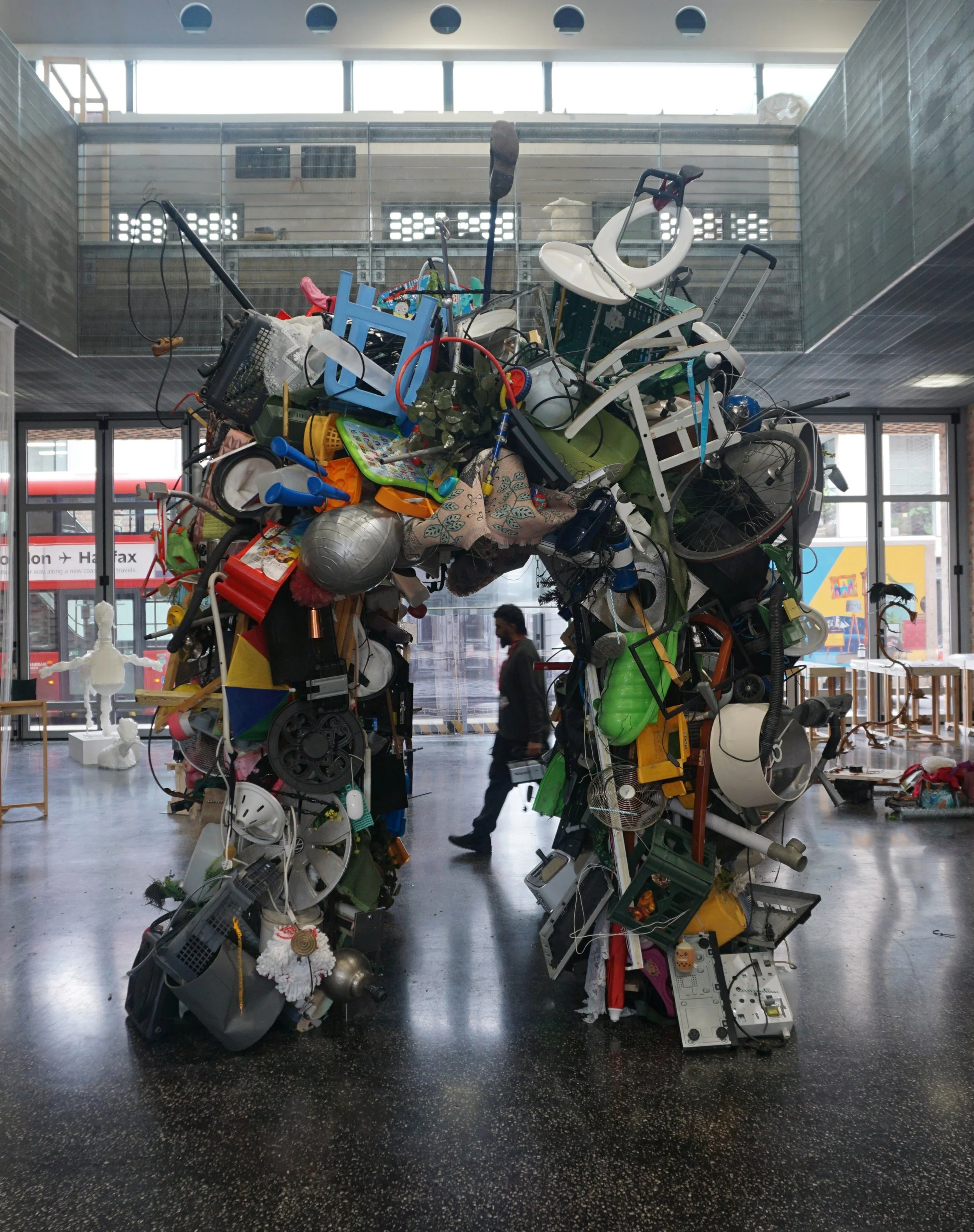 A large art sculpture made of discarded household objects and appliances, shaped like a human figure, standing indoors in a building with large windows.