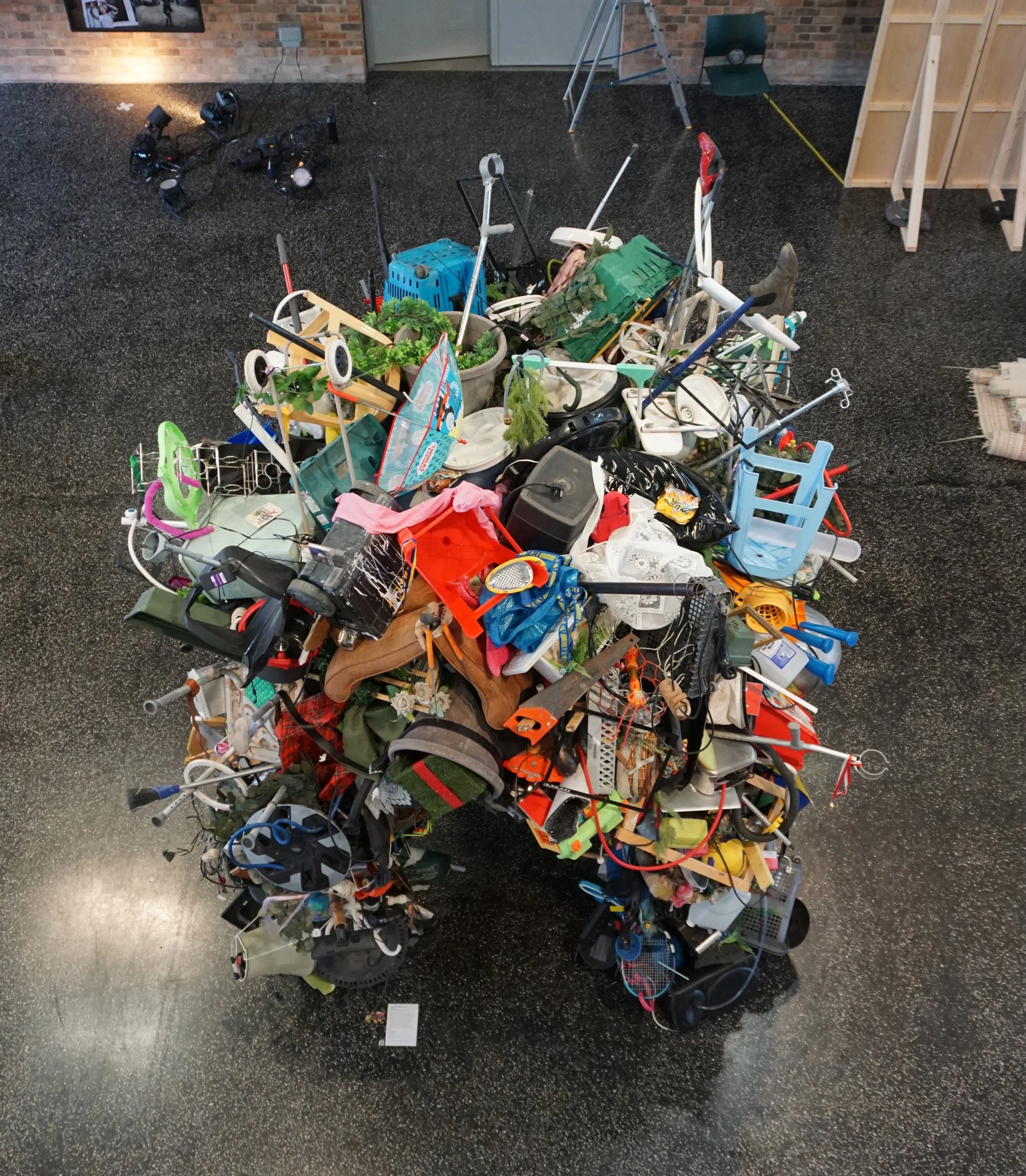 A large pile of assorted used children's toys and household items on an indoor black speckled floor, including bicycles, a tricycle, a shopping cart, gardening tools, a plastic crate, and various other objects.
