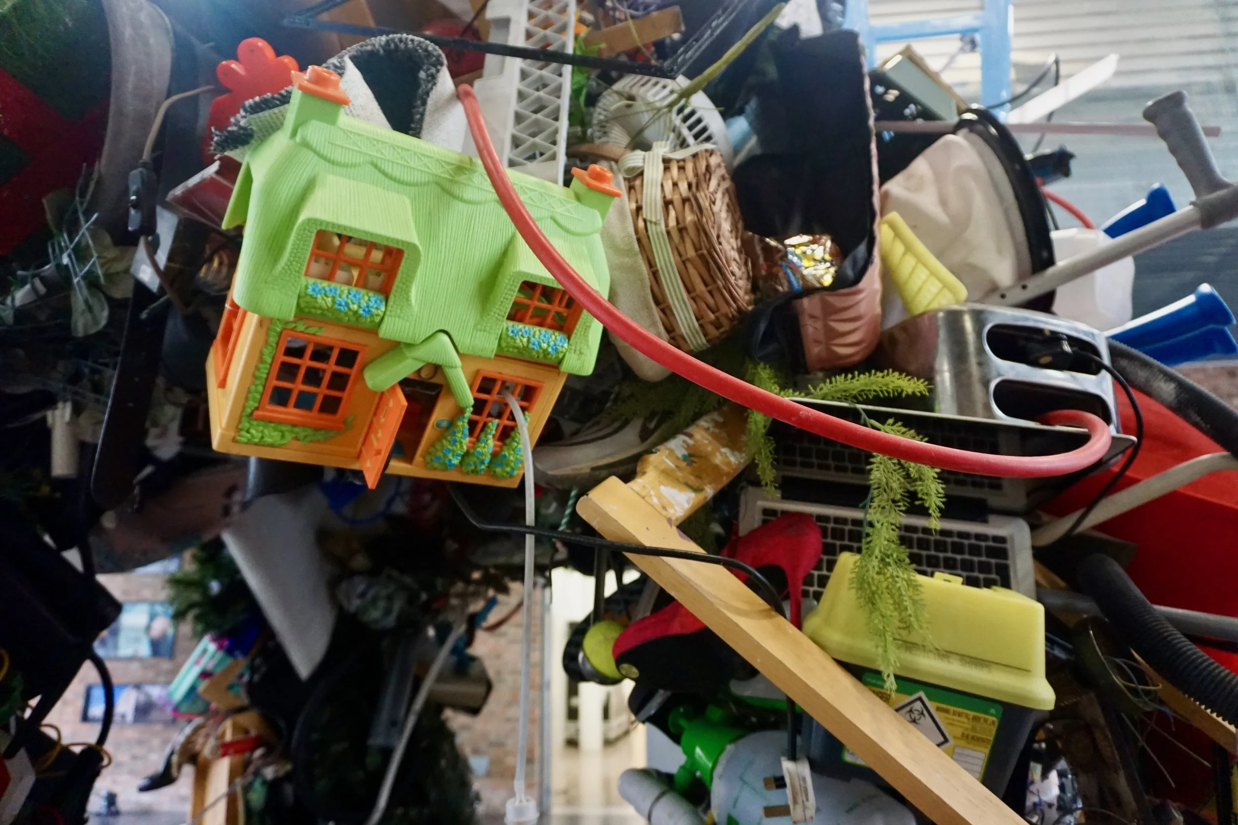 A cluttered store shelf filled with miscellaneous items including a small green toy house, baskets, tools, and household objects.