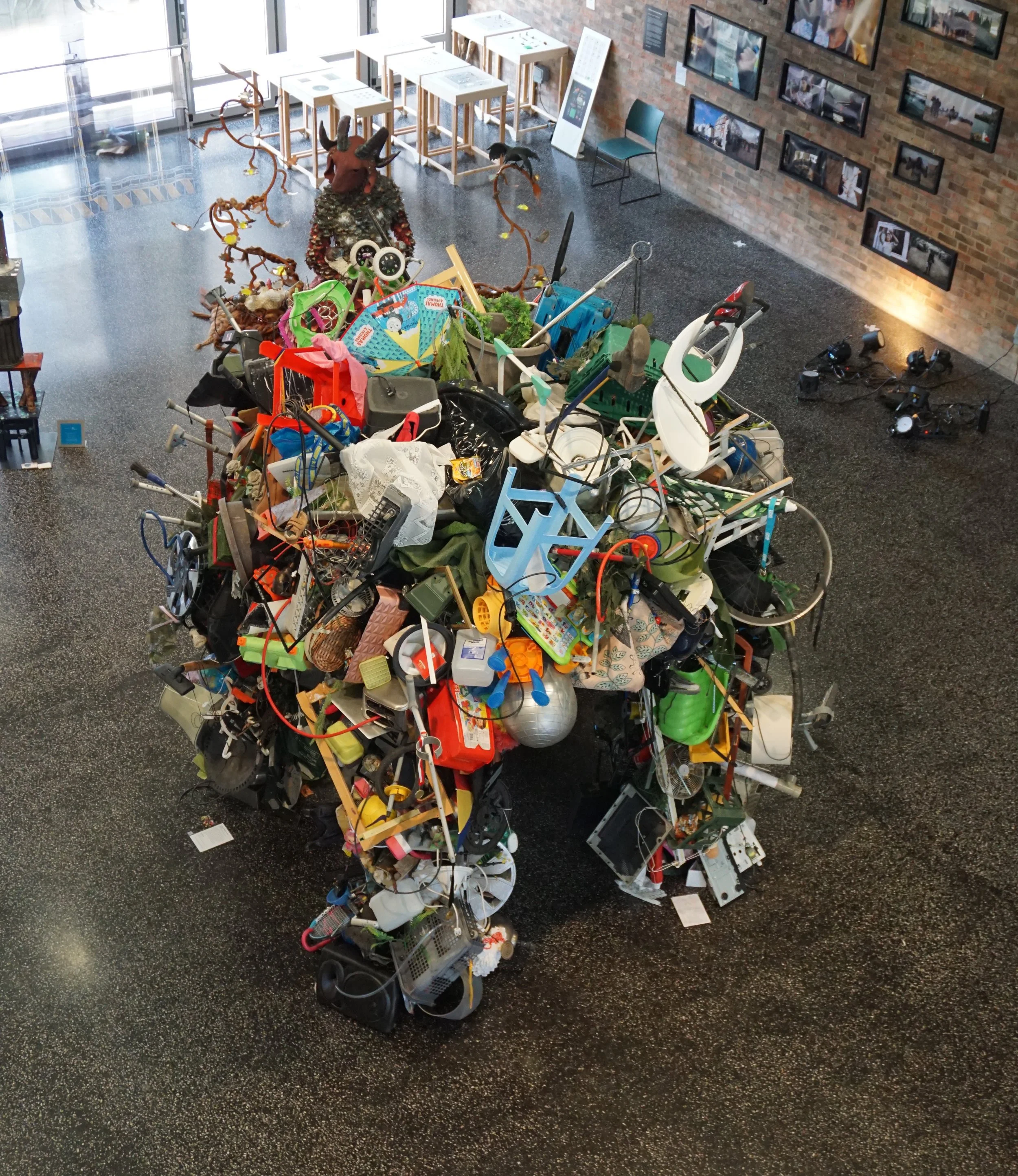 A large pile of used and discarded household items and trash inside a gallery or exhibit space, with a brick wall on one side and framed photographs on the wall, and a chair visible nearby.