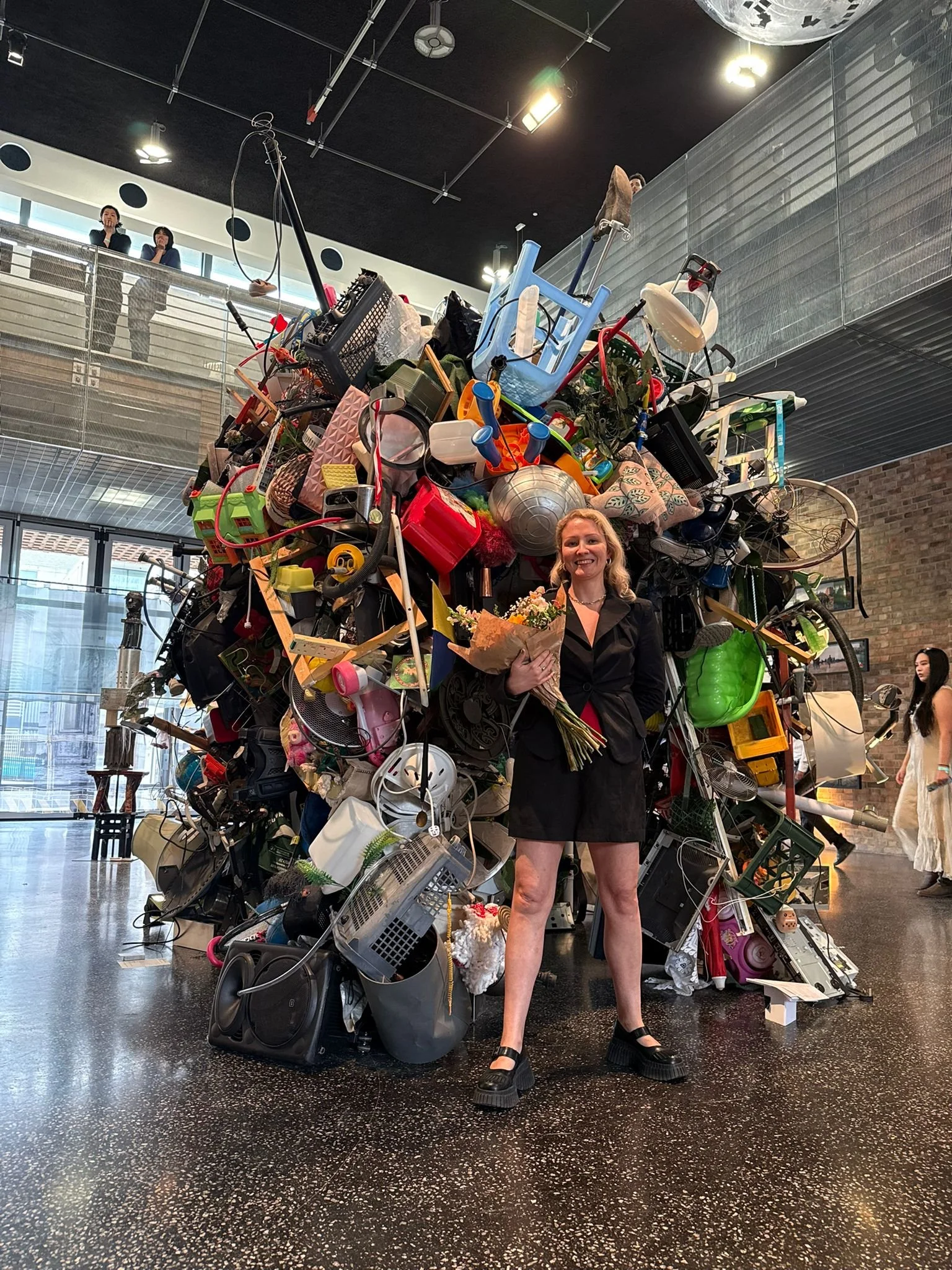 A woman standing in front of a large sculpture made of assorted discarded objects, holding a bouquet of flowers and smiling. The sculpture is an abstract form constructed from various household items like kitchenware, electronic parts, and toys, displayed indoor with a polished black floor and modern interior design.