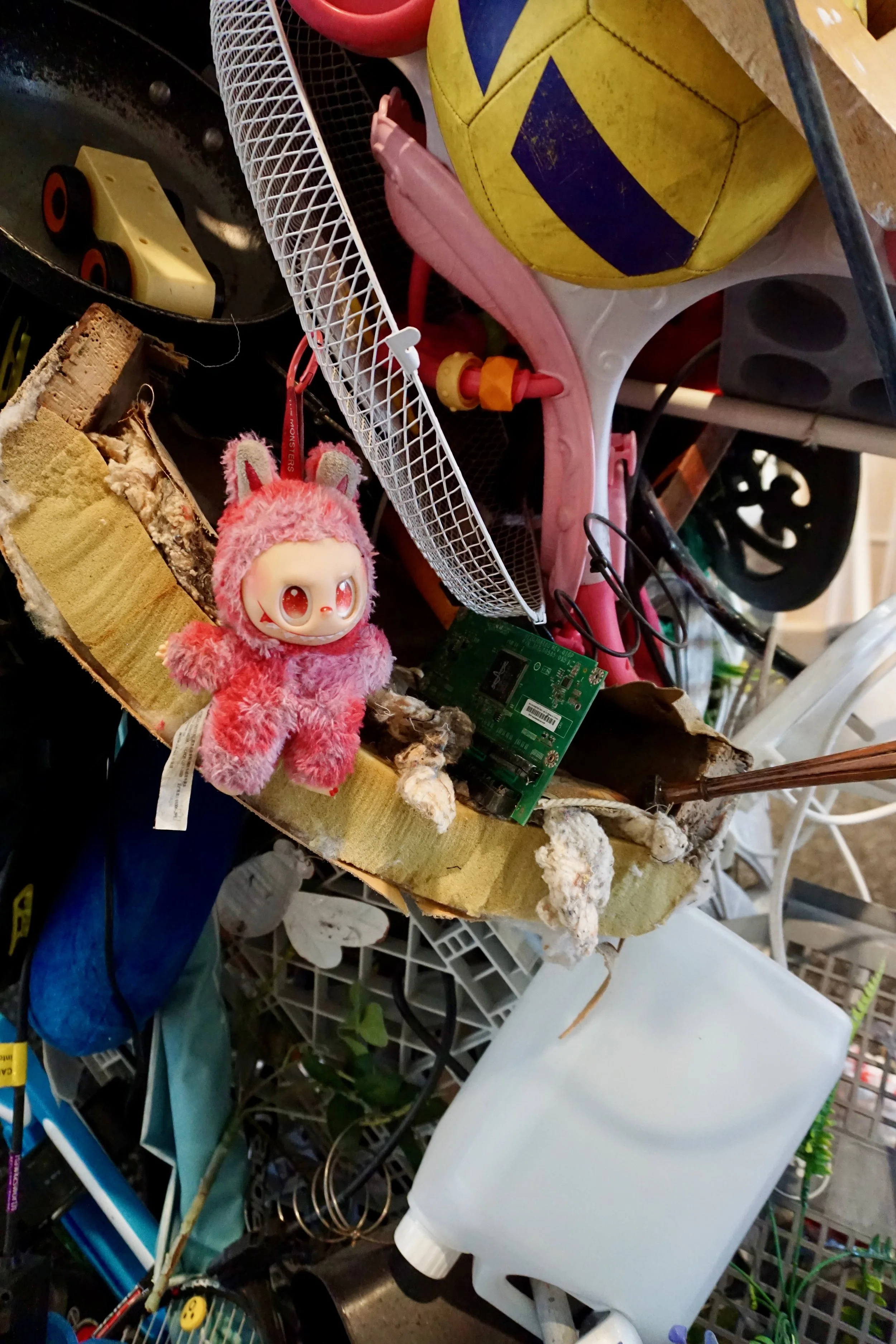 A cluttered table with a yellow and purple basketball, a pink plush toy, a white plastic bottle, and various other objects including a circuit board, wood pieces, and a rake. The background includes metal wire shelves and various tools and items.