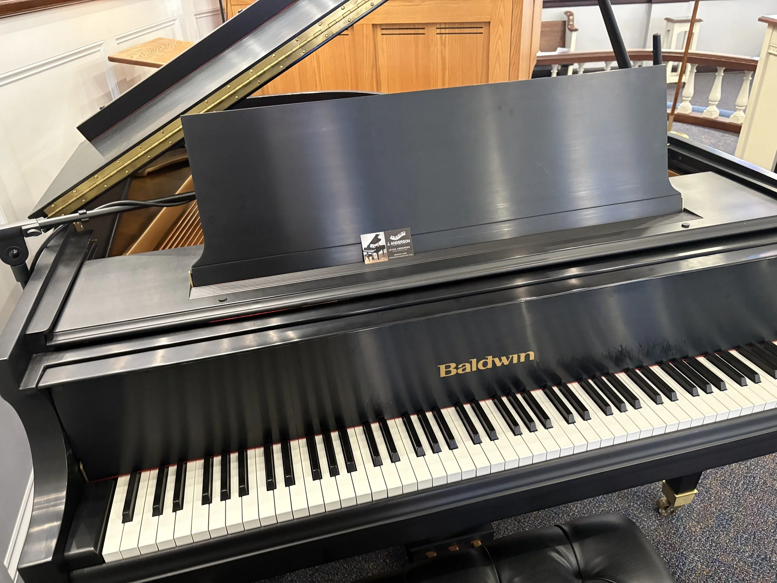 A Baldwin grand piano with a black finish, open lid, and a music stand, situated in a room with wooden paneling and a carpeted floor.