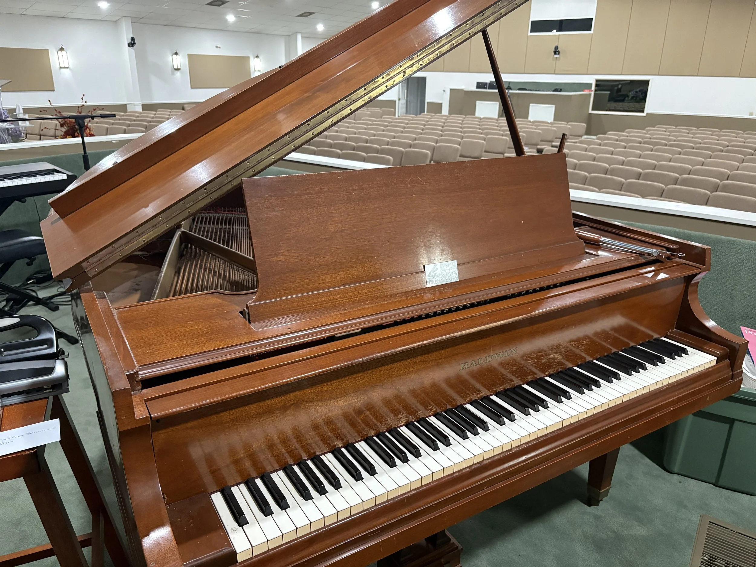 A wooden Baldwin grand piano in a large room with beige seating and a green carpet. The piano's lid is open, exposing the strings and inner mechanics.