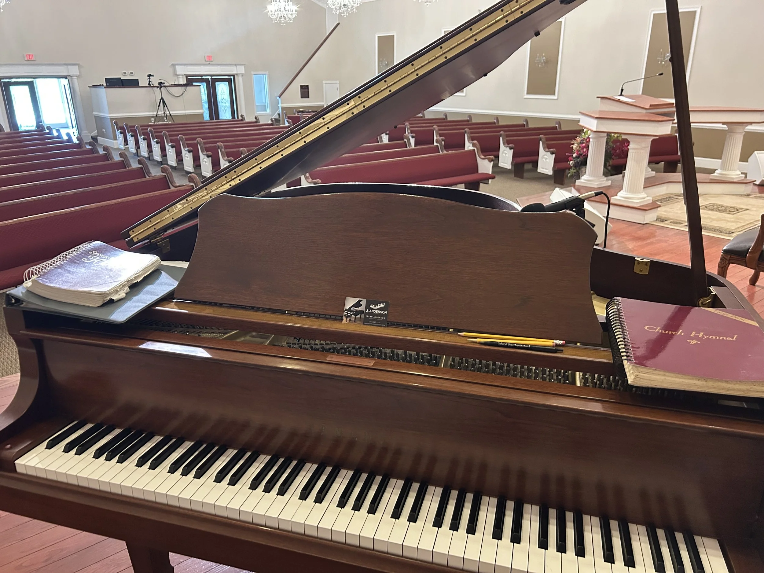 A grand piano inside a church sanctuary with red pews, a pulpit, and floral arrangements in the background.