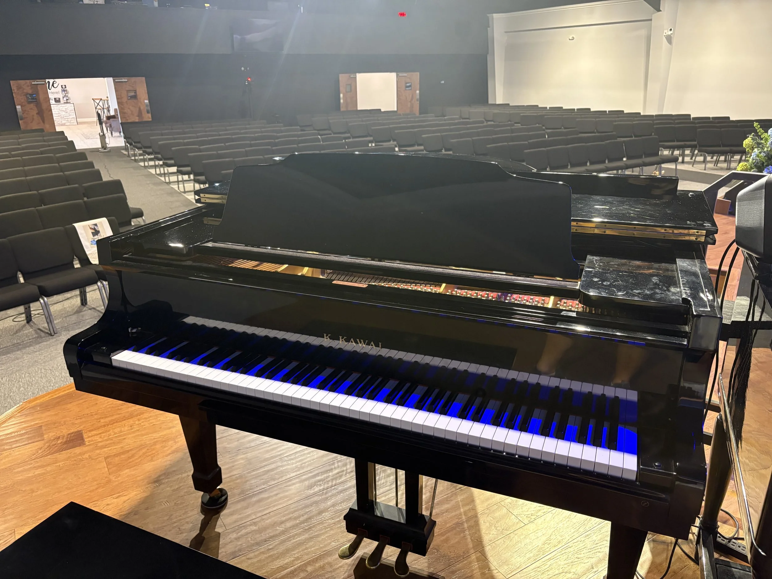 A black grand piano on a wooden stage in an auditorium or event hall with multiple rows of chairs and doors in the background.