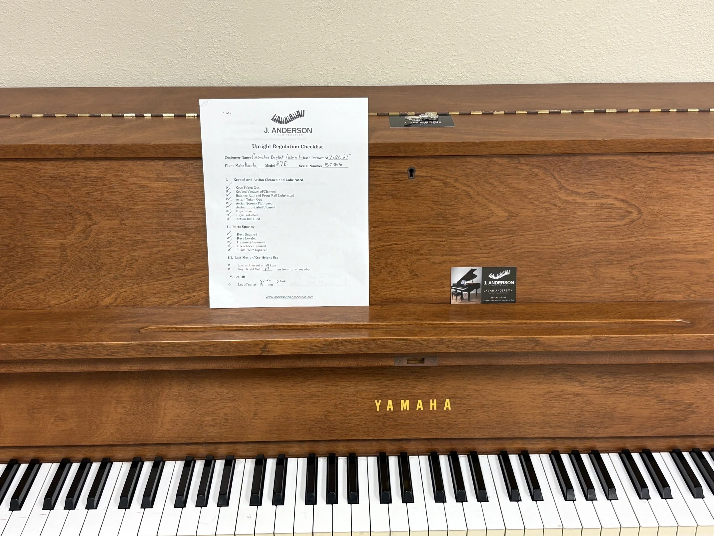 A Yamaha upright piano with a wood finish, a regulation checklist paper on top, and a small business card below the paper on the right side, against a beige wall.