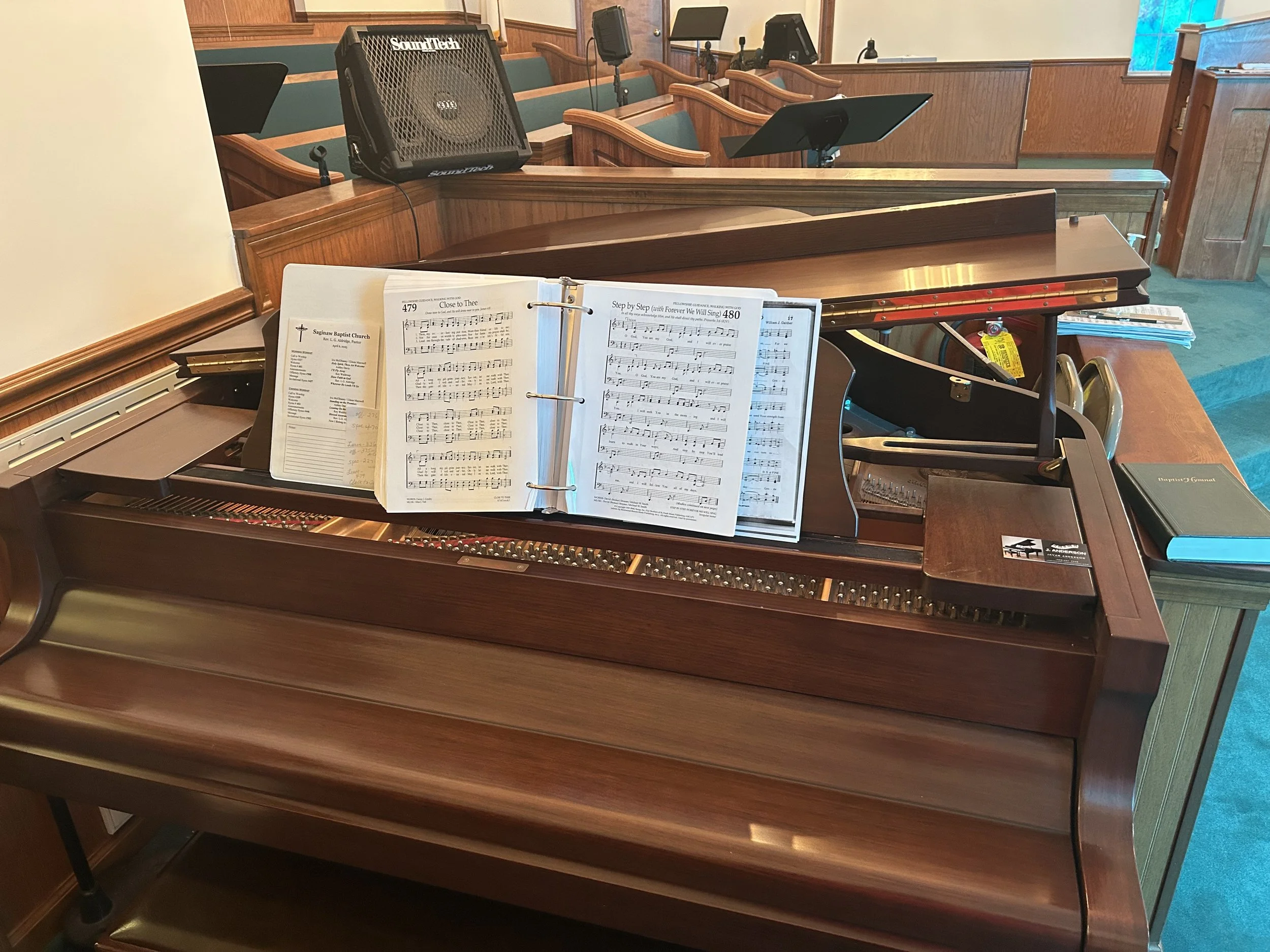 A wooden church organ with open sheet music and hymnals, located in a church sanctuary with pews and music stands in the background.