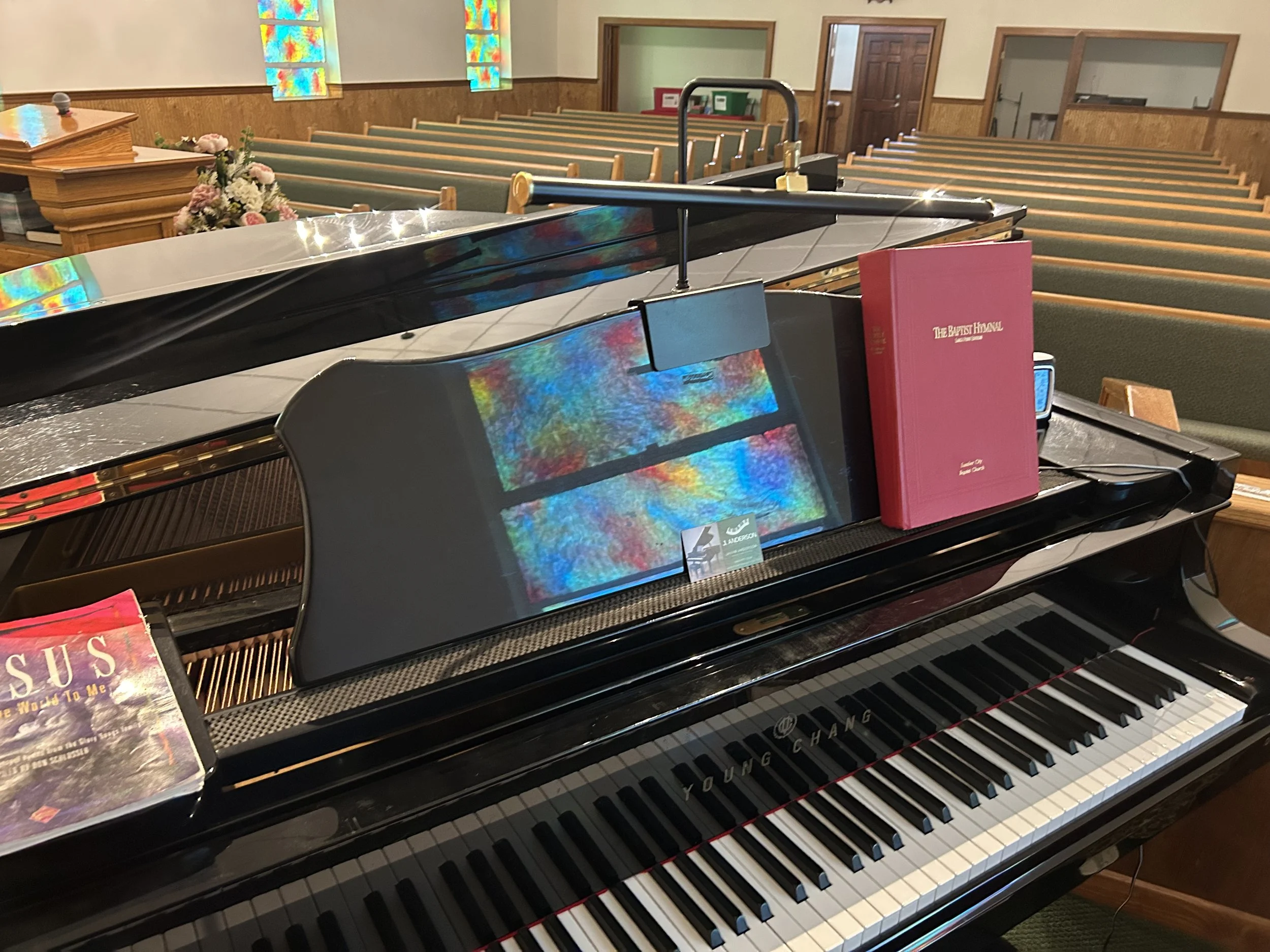 A black Yamaha grand piano with books on top, inside a church with wooden pews, stained glass windows, and flowers.
