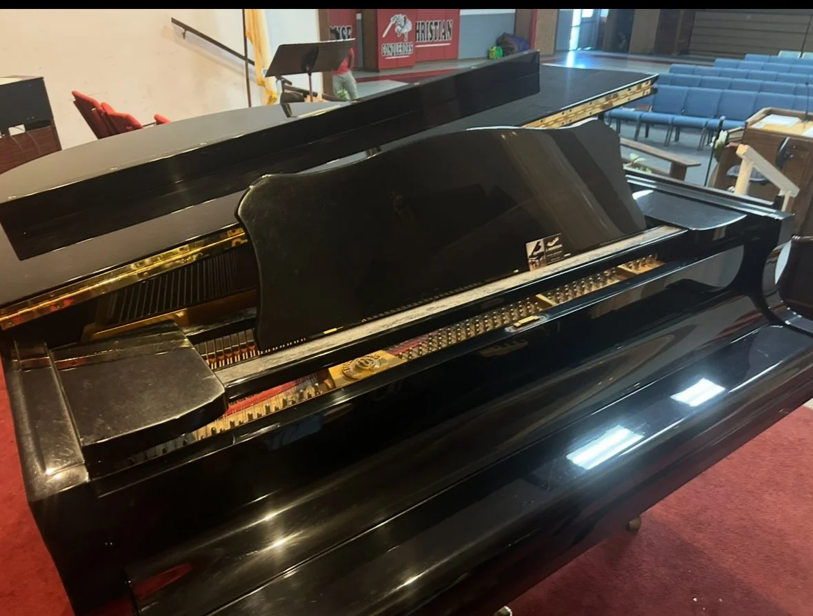 A black grand piano with its lid open, revealing the strings and internal components, situated in a spacious room with chairs and banners in the background.