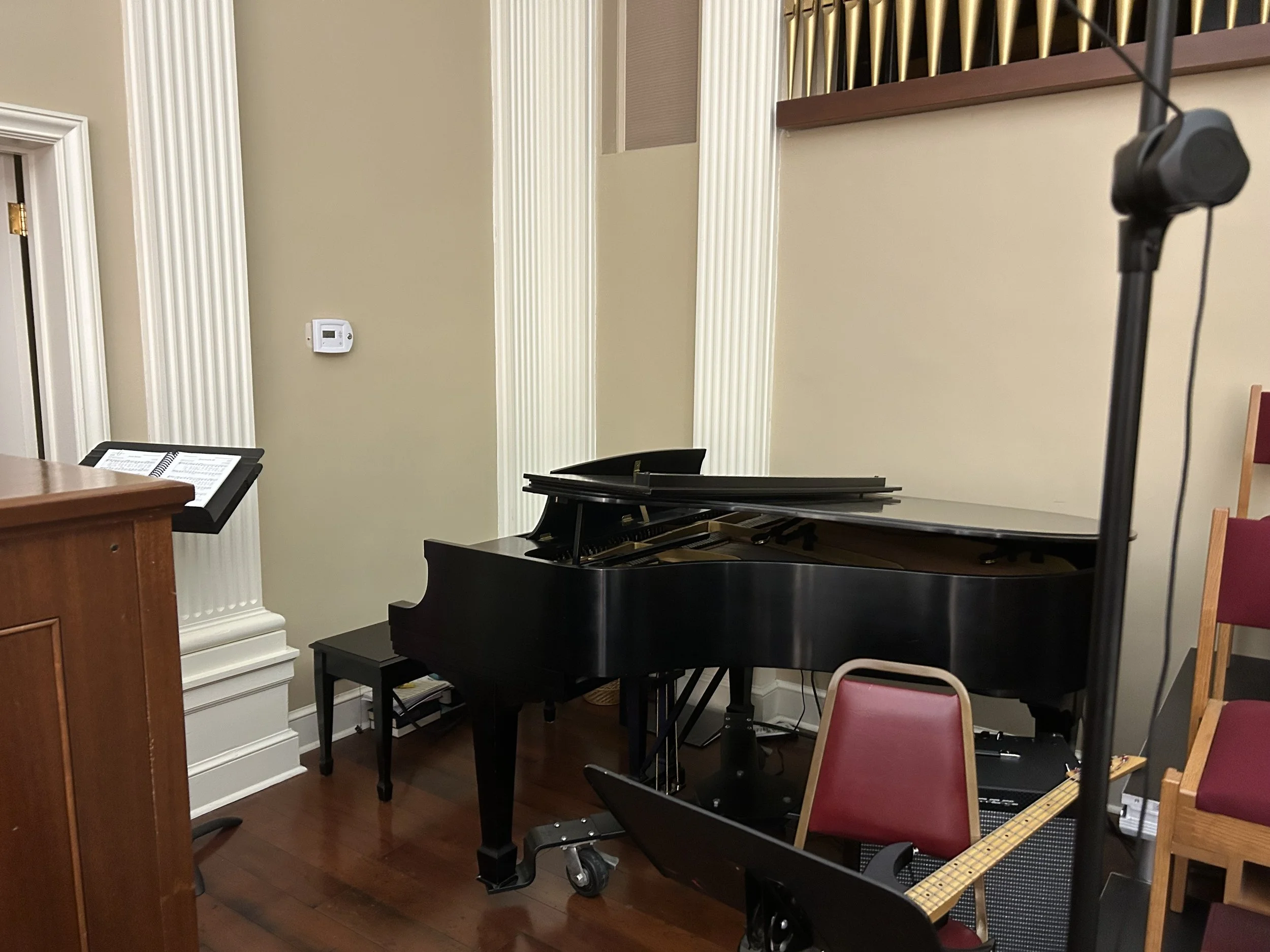 A living room with a grand piano, a music stand with sheet music, a guitar on a stand, and chairs against the wall.