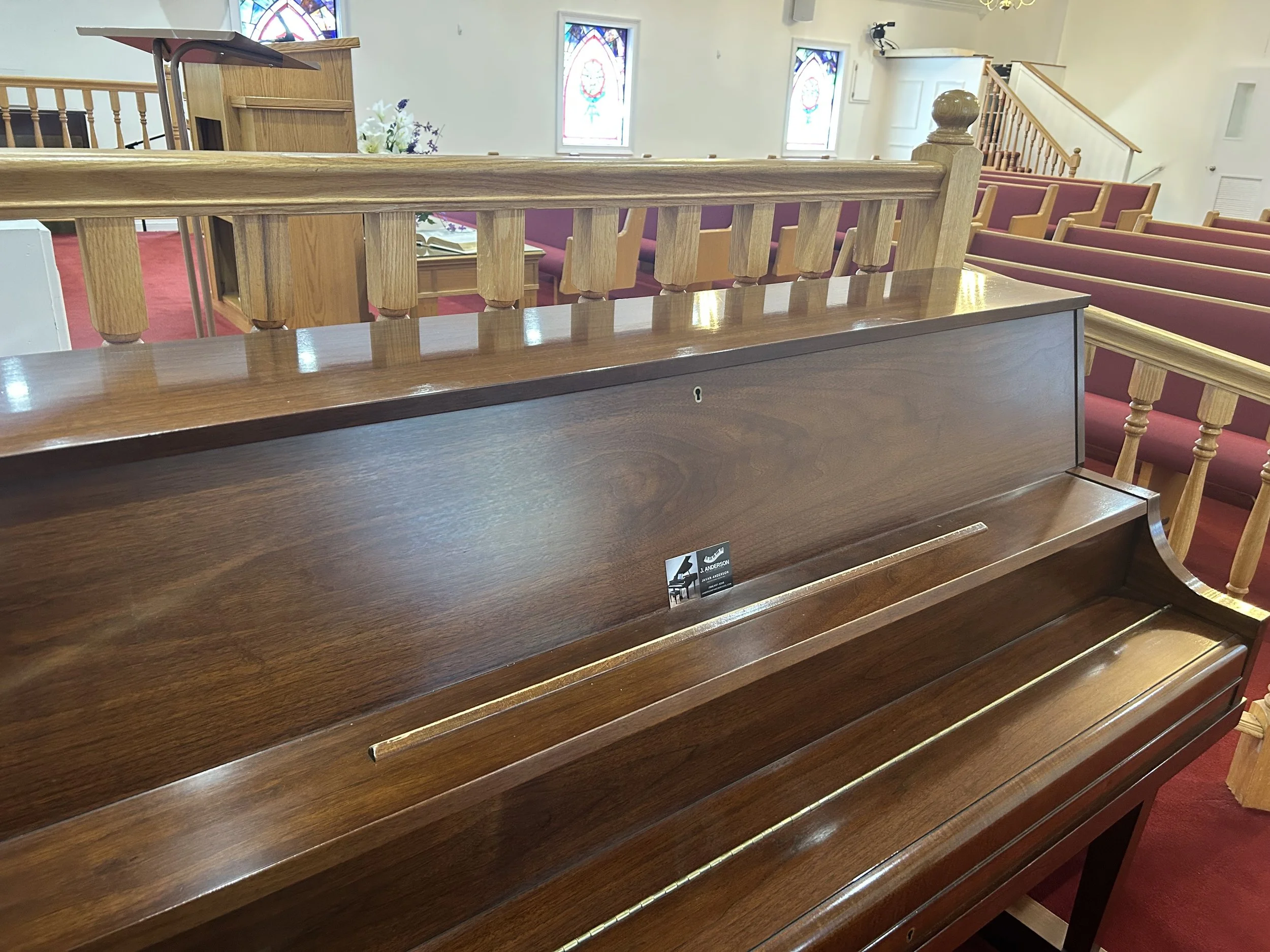 Wooden piano in a church with stained glass windows and red pews.
