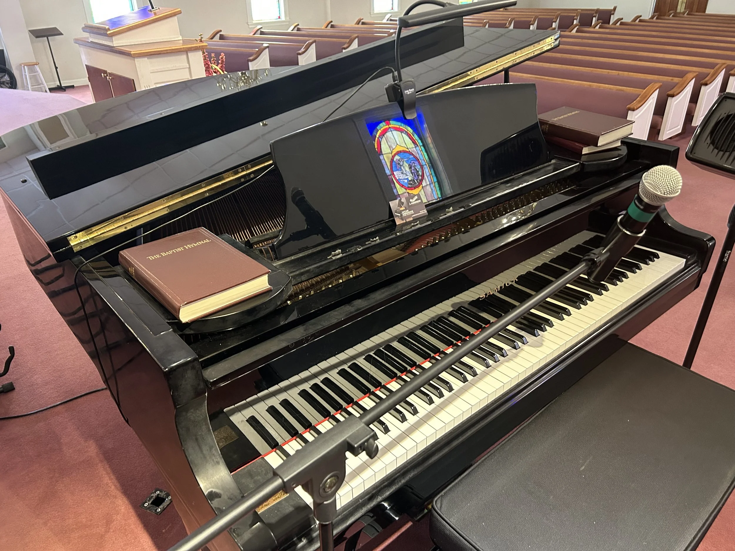 A black grand piano with a microphone in front of it and a hymnal book on the left side, located inside a church with empty pews and stained glass windows.