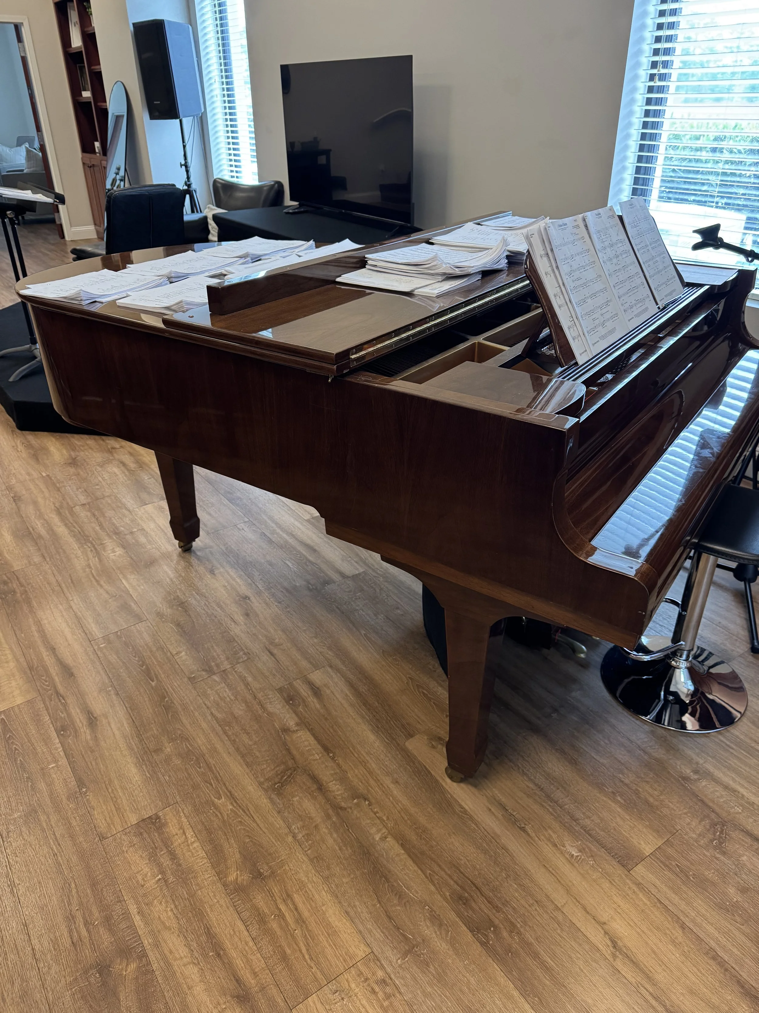 A wooden grand piano with open sheet music on the music stand, located in a room with wood flooring, a TV, window blinds, and some furniture.