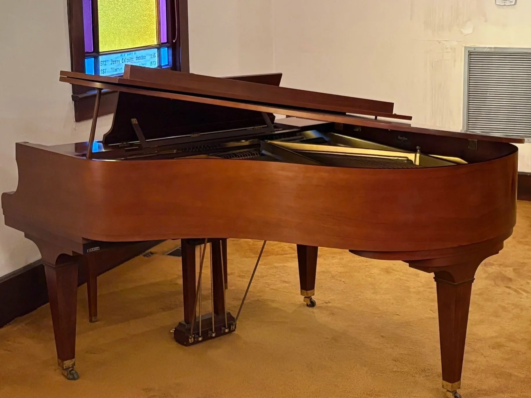 A brown grand piano with a closed lid in a room with wooden flooring and a small stained glass window.