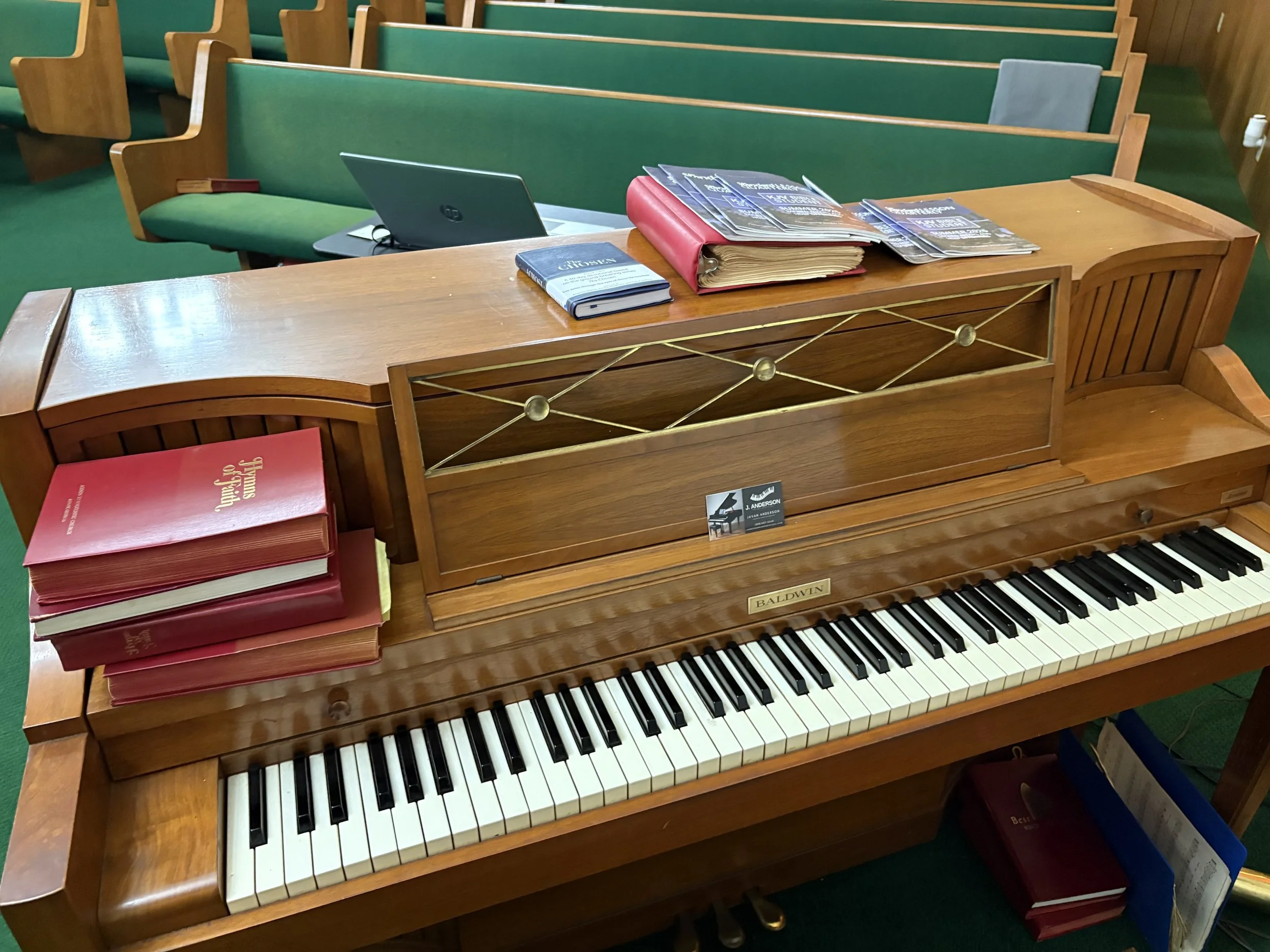 An upright Baldwin piano with books and sheet music on top, located in a room with green carpet and wooden pews.