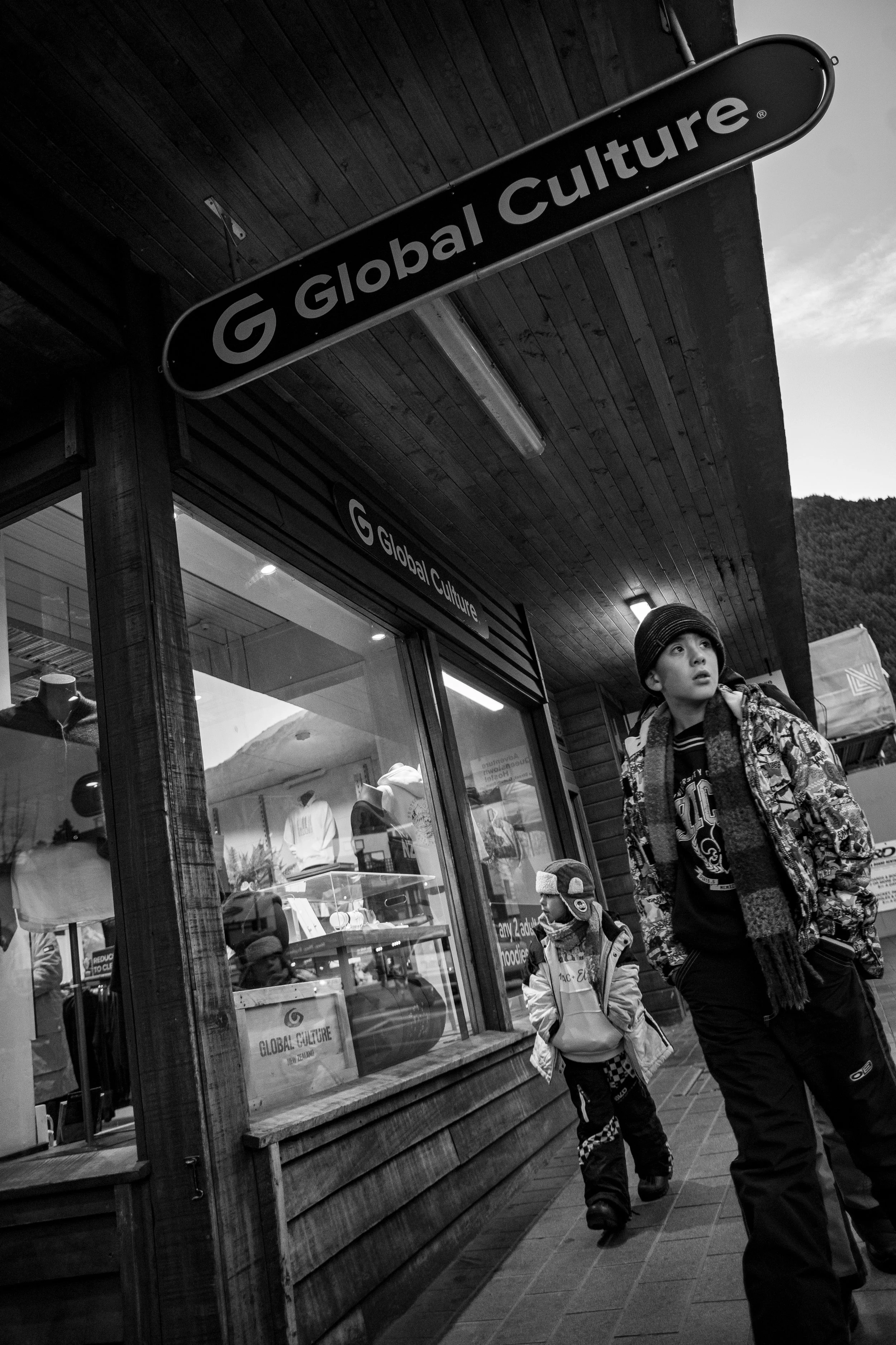 A black and white photo of a storefront with a sign that reads 'Global Culture' and two young boys walking past, dressed in winter clothing, with mountains in the background.