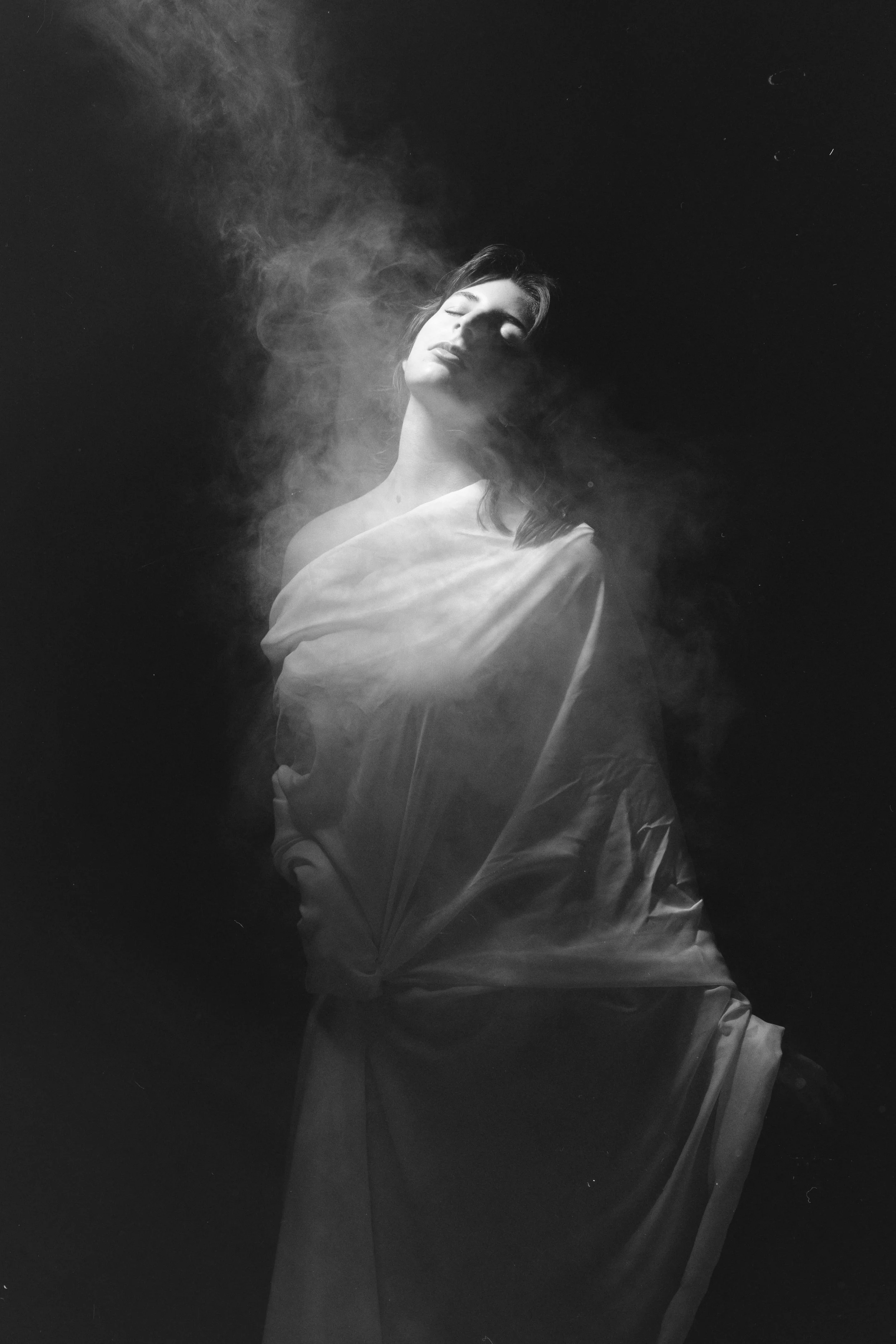 A black and white photograph of a woman with eyes closed, wrapped in a sheer cloth with smoke or mist surrounding her, creating a dramatic and ethereal atmosphere.