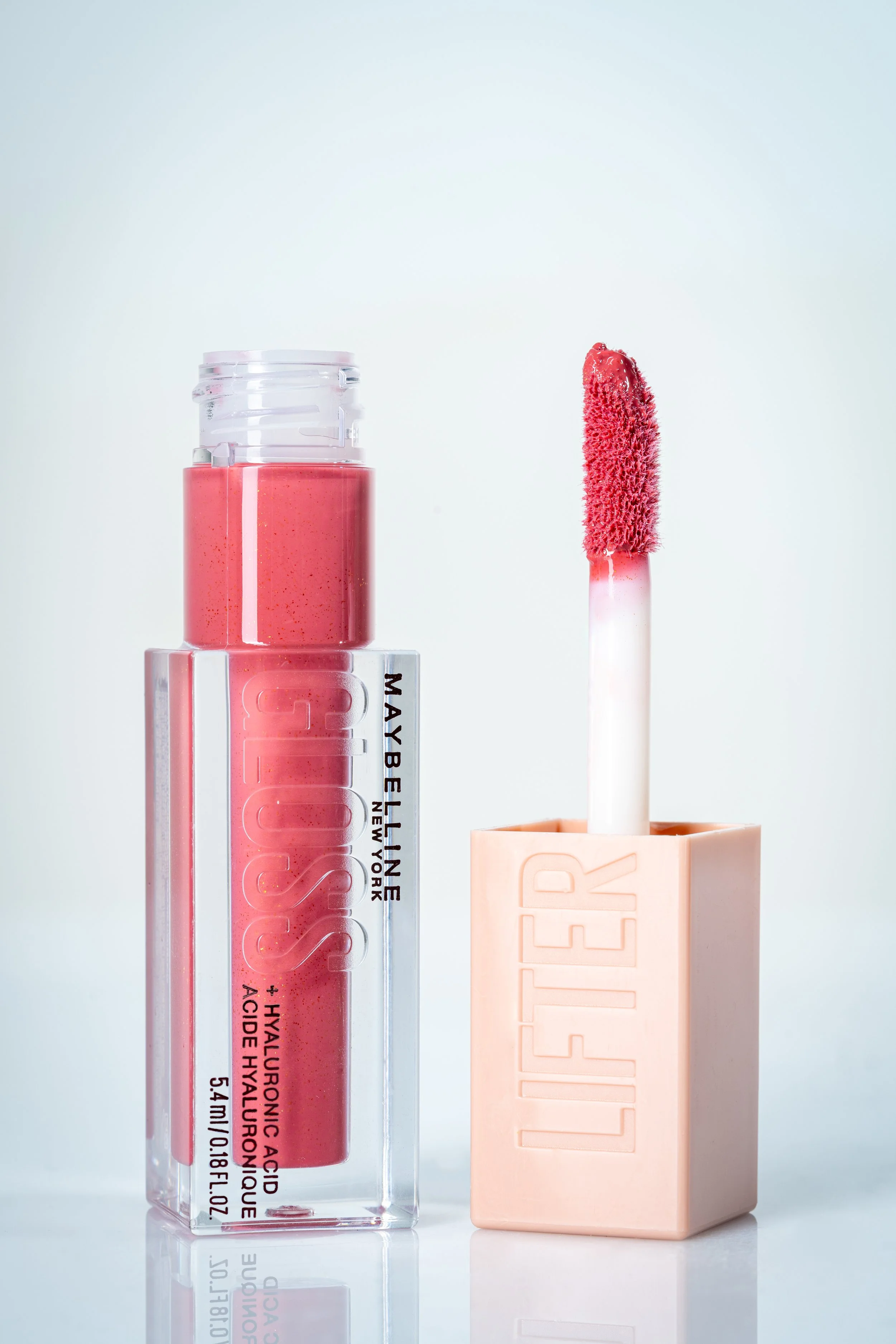 A Maybelline lip gloss tube and a lip gloss applicator with a pink lip gloss inside. The tube has "LIPSTICK" embossed on it, and the applicator has a pink sponge tip.