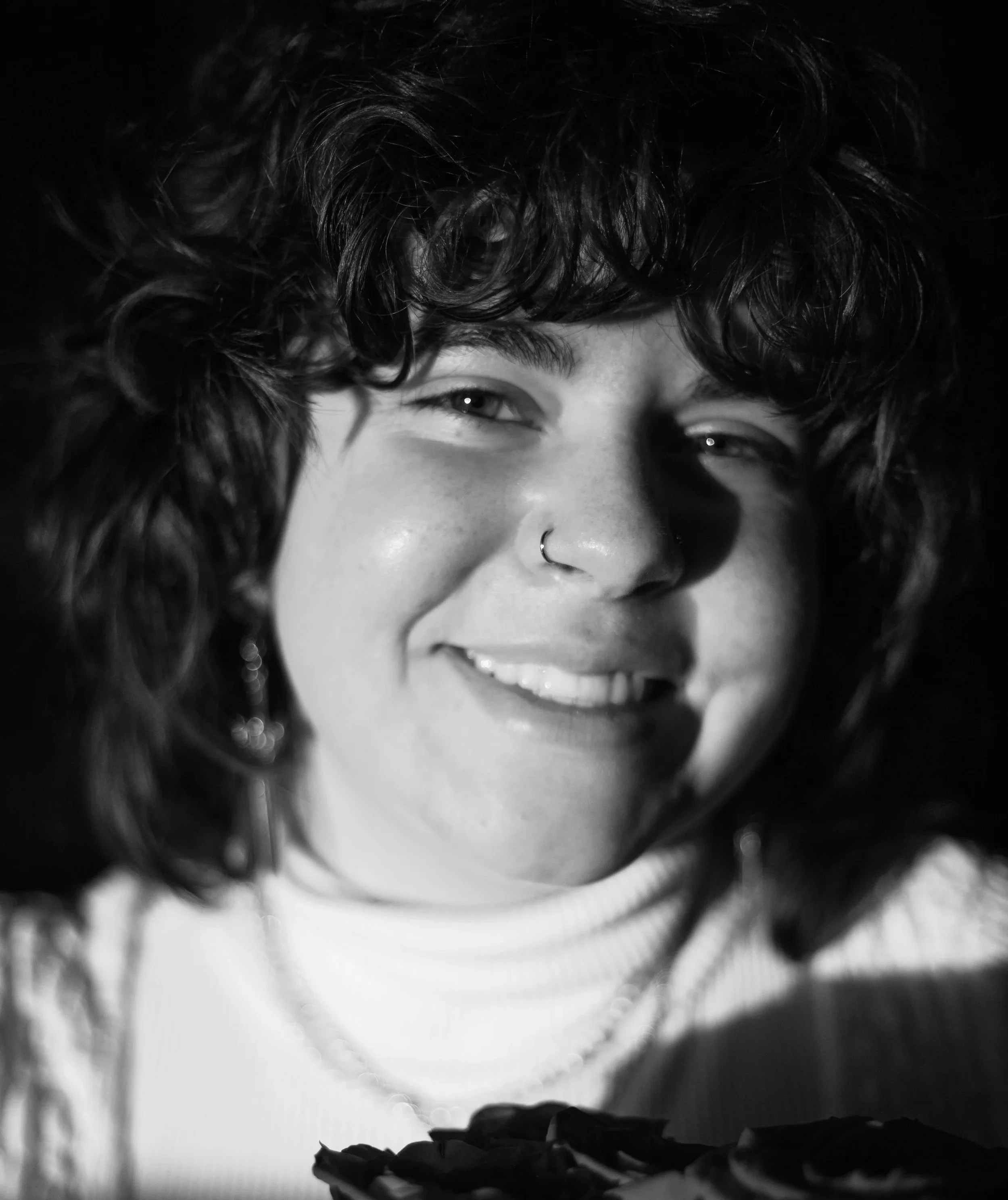 A black and white close-up photo of a young woman with curly hair smiling at the camera, wearing a nose ring, earrings, and a turtleneck sweater.