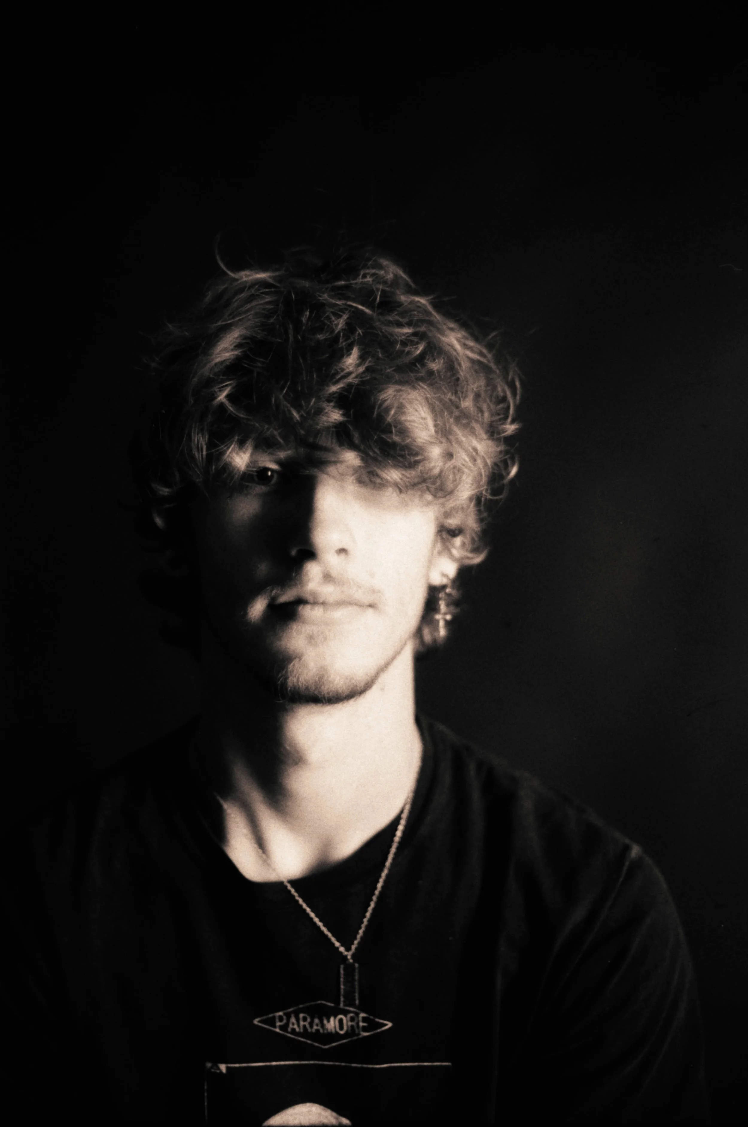 Black and white portrait of a young man with curly hair wearing a Paramore t-shirt, looking at the camera with a neutral expression.