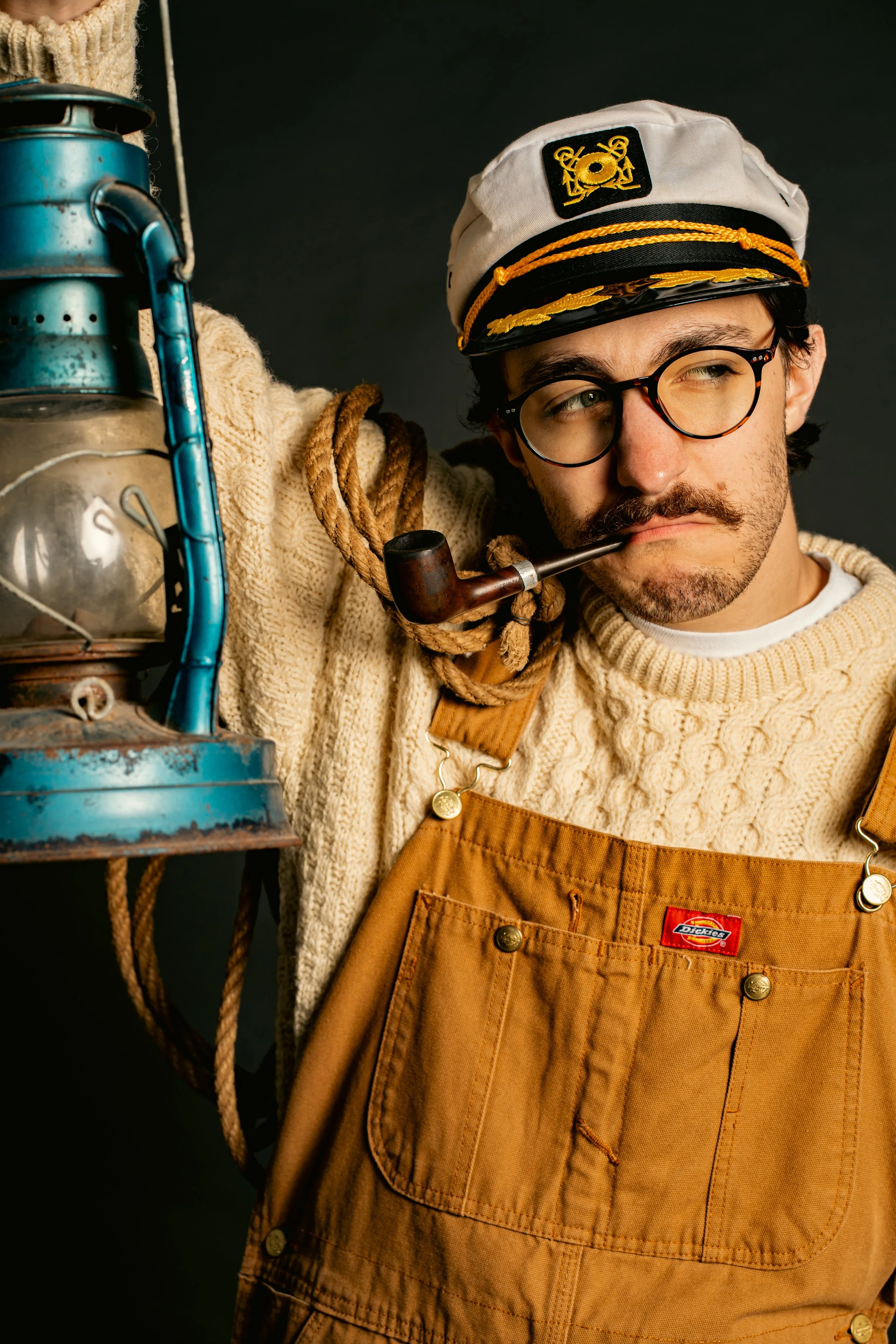 A man dressed as a sailor with glasses, a pipe, and a captain's hat, wearing a knitted sweater and brown overalls, holding a vintage lantern and a rope.