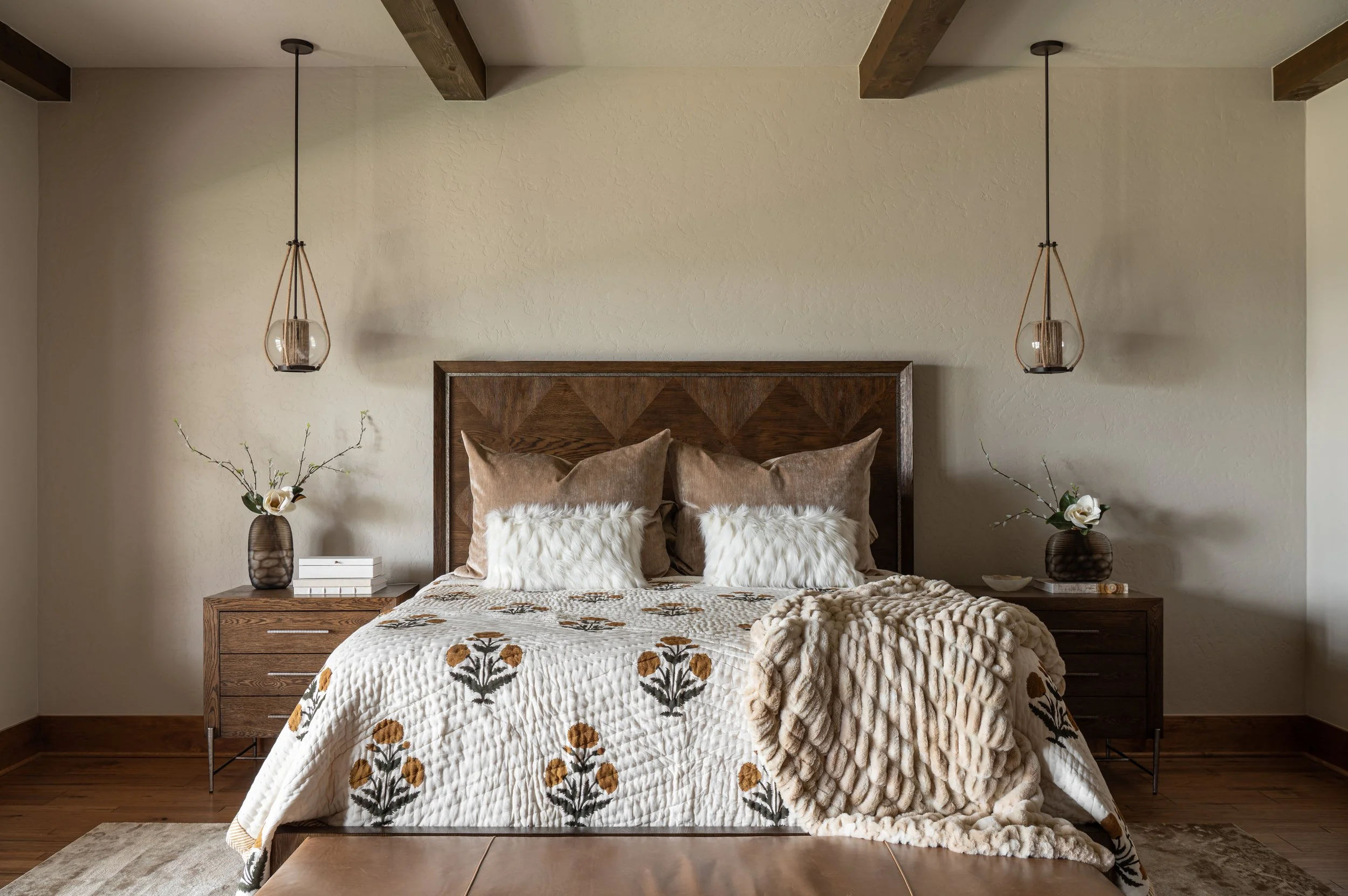 A neatly made bed with a wooden headboard, beige pillows, and a floral quilt. Two nightstands with decorative vases holding flowers are on either side, with pendant lights hanging from the ceiling.
