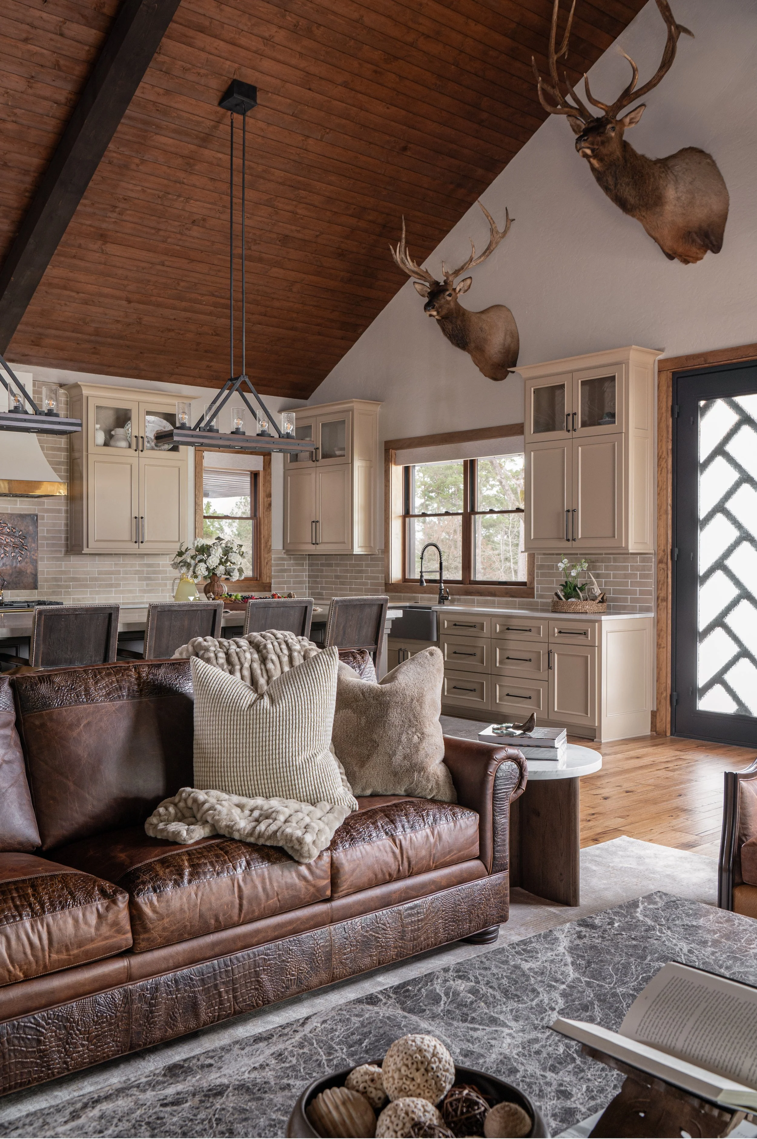 Living room with a leather sofa, decorative pillows, a marble coffee table with a bowl of decorative balls, a kitchen area with cream cabinets, a window, and mounted deer heads on the wall under a wooden vaulted ceiling.