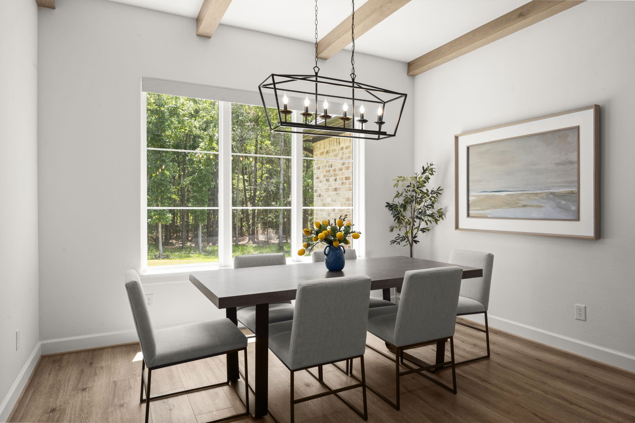 A dining room with a rectangular wooden table, six light gray upholstered chairs, a blue vase with yellow flowers, and a large window showing a green outdoor area.