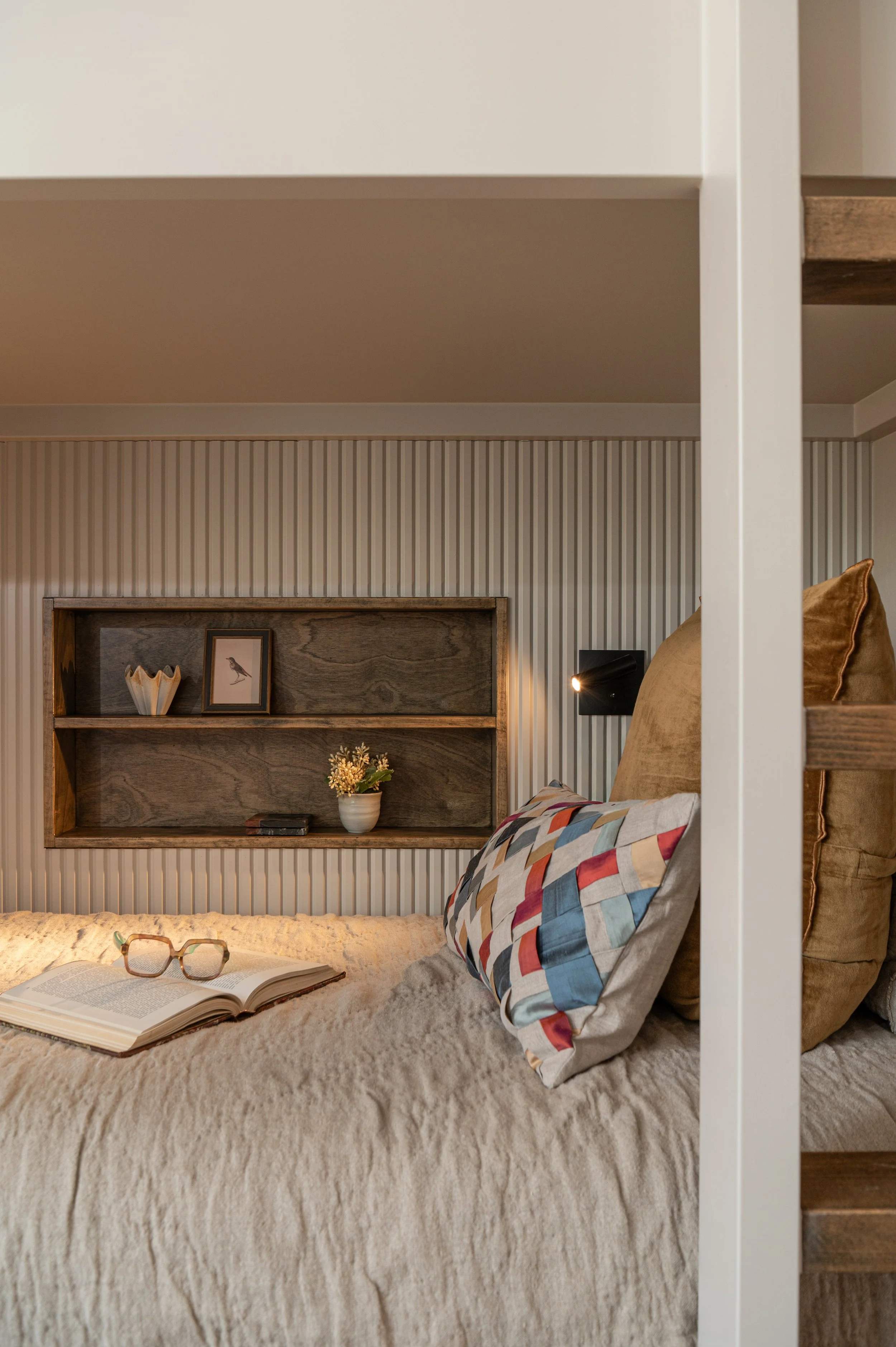 A cozy bedroom scene with a beige textured bedspread, a pair of glasses, and an open book. The bed has multi-colored geometric pillows. Behind the bed, there is a wall with vertical striped wallpaper, and a wooden shelf with decorative items, includi
