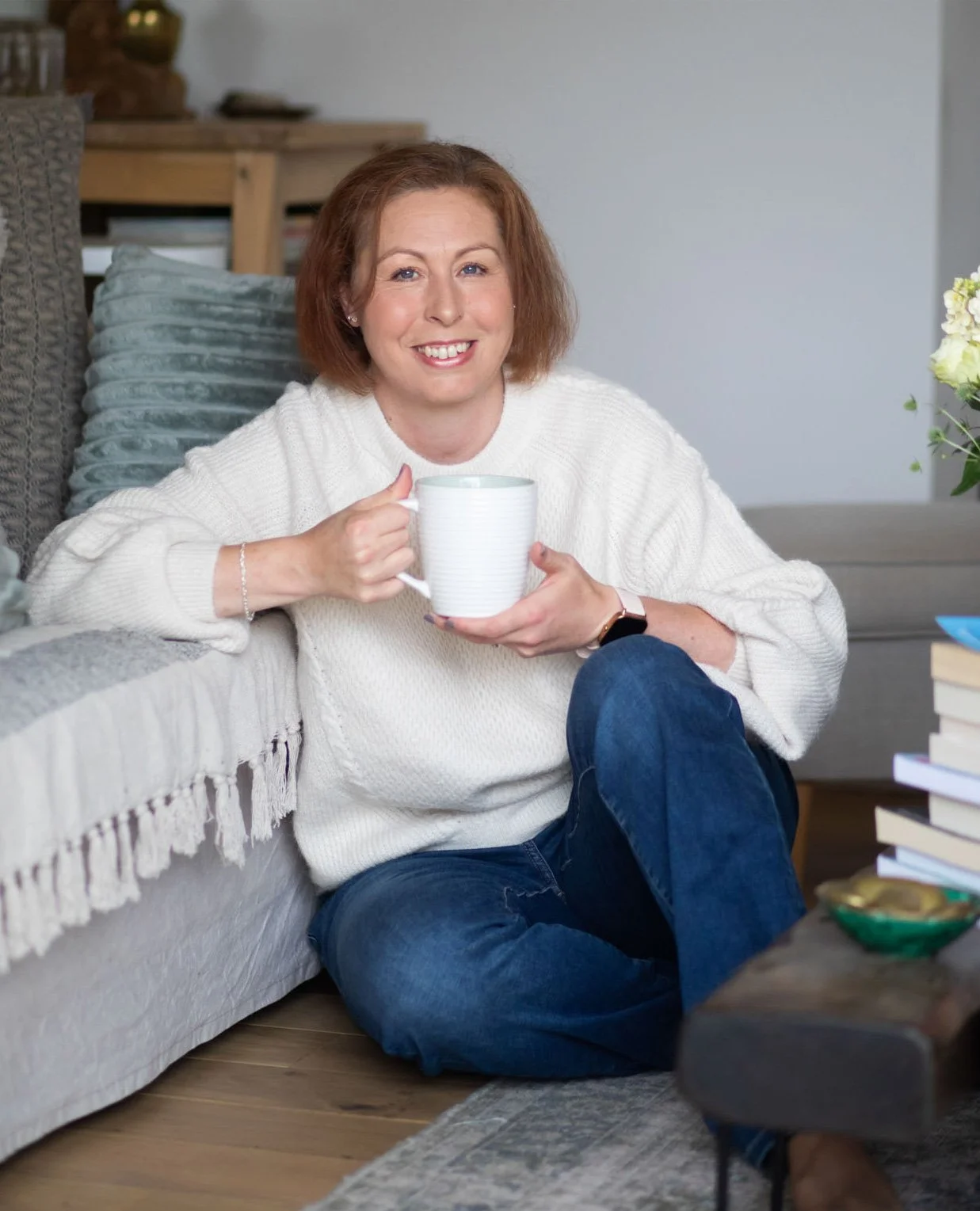 A woman, wearing a white sweater and jeans, sitting on the floor next to a sofa, holding a white mug and smiling at the camera in a cozy living room.