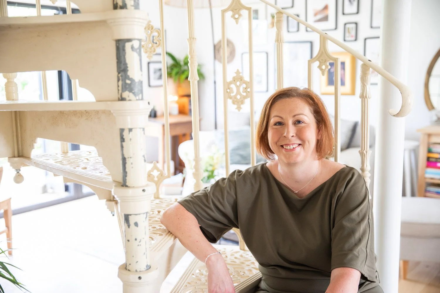 A woman with short  hair and a green top sitting on a staircase inside a well-lit living room, smiling at the camera.