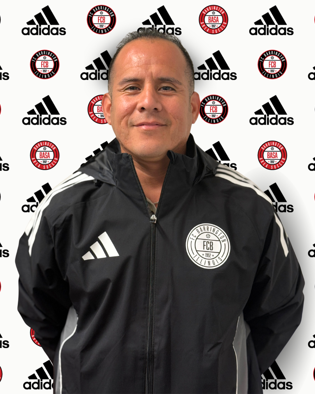 A man standing in front of a backdrop with Adidas and Barrington Area Soccer logos, wearing a gray Adidas jacket with a FCX logo.