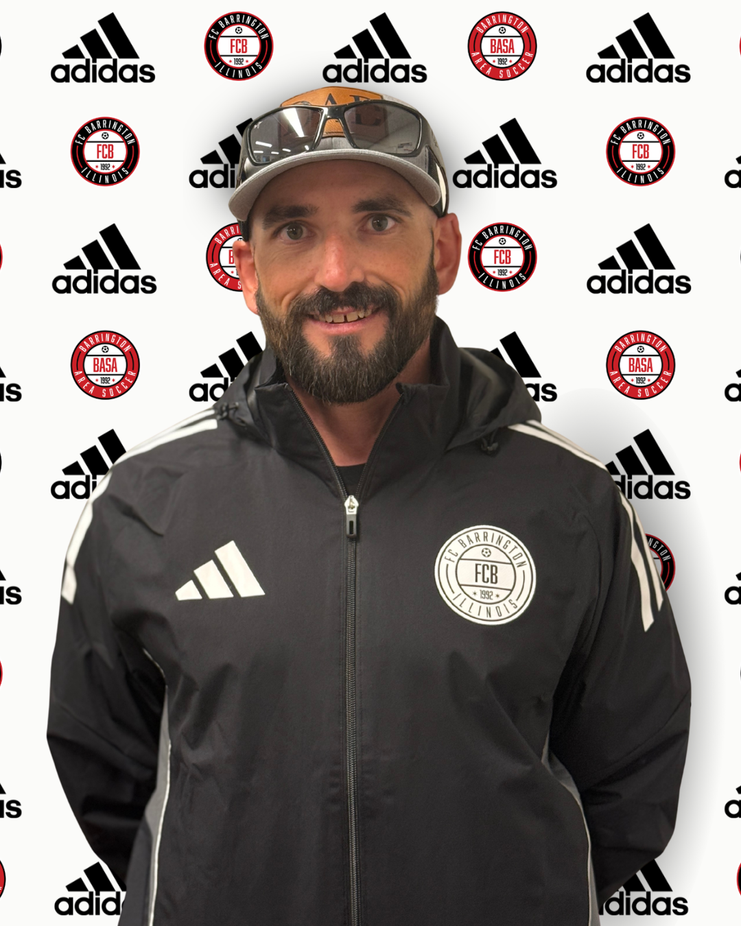 Man in gray Adidas jacket and gray baseball cap standing in front of a backdrop with Adidas and Barrington Area Soccer logos.