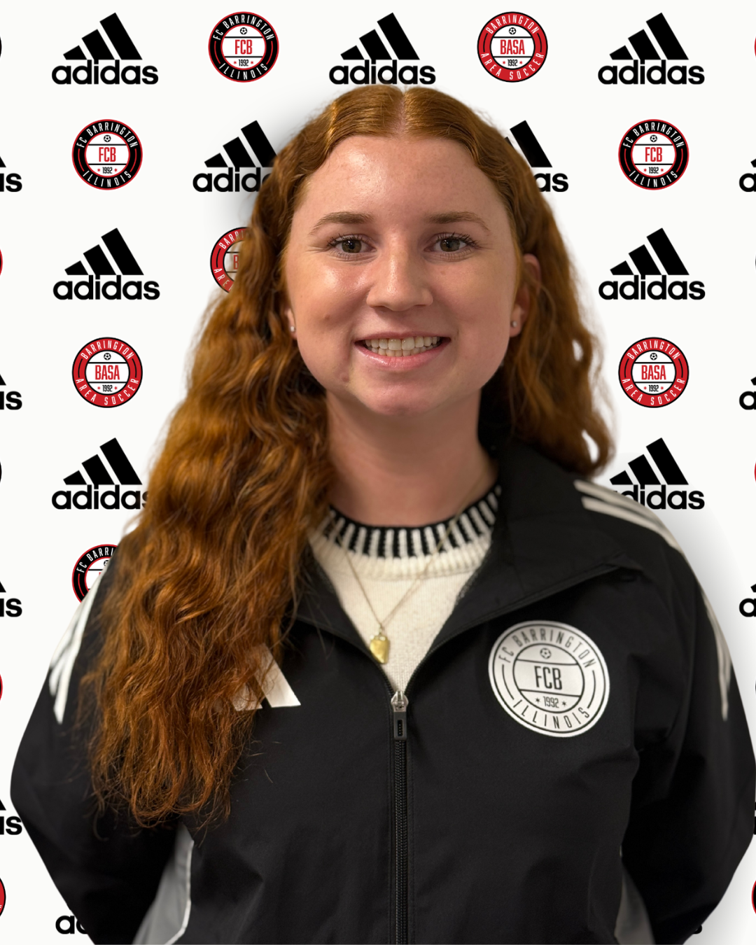 A woman wearing a gray Adidas jacket and black t-shirt standing in front of a white backdrop with repeated logos of Adidas, FC Barrington Illinois, and Barrington Area Soccer.