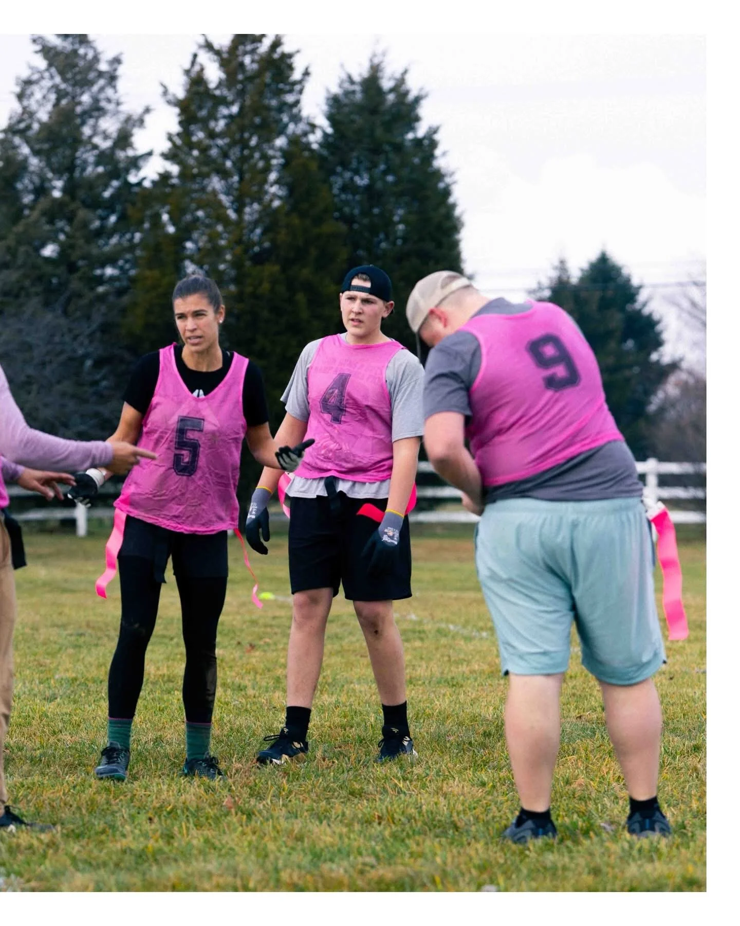 Group of people wearing pink sports pinnies standing on a grassy field, with trees in the background.