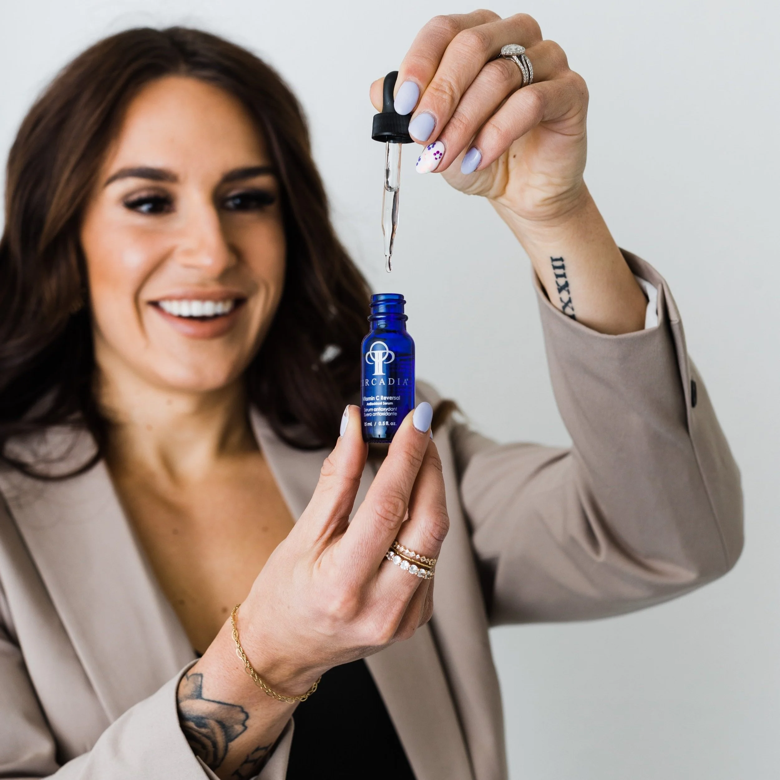 A woman with brown hair and tattoos on her arm is smiling while holding a blue bottle of serum and a dropper, with a drop of serum about to fall from the dropper into the bottle.