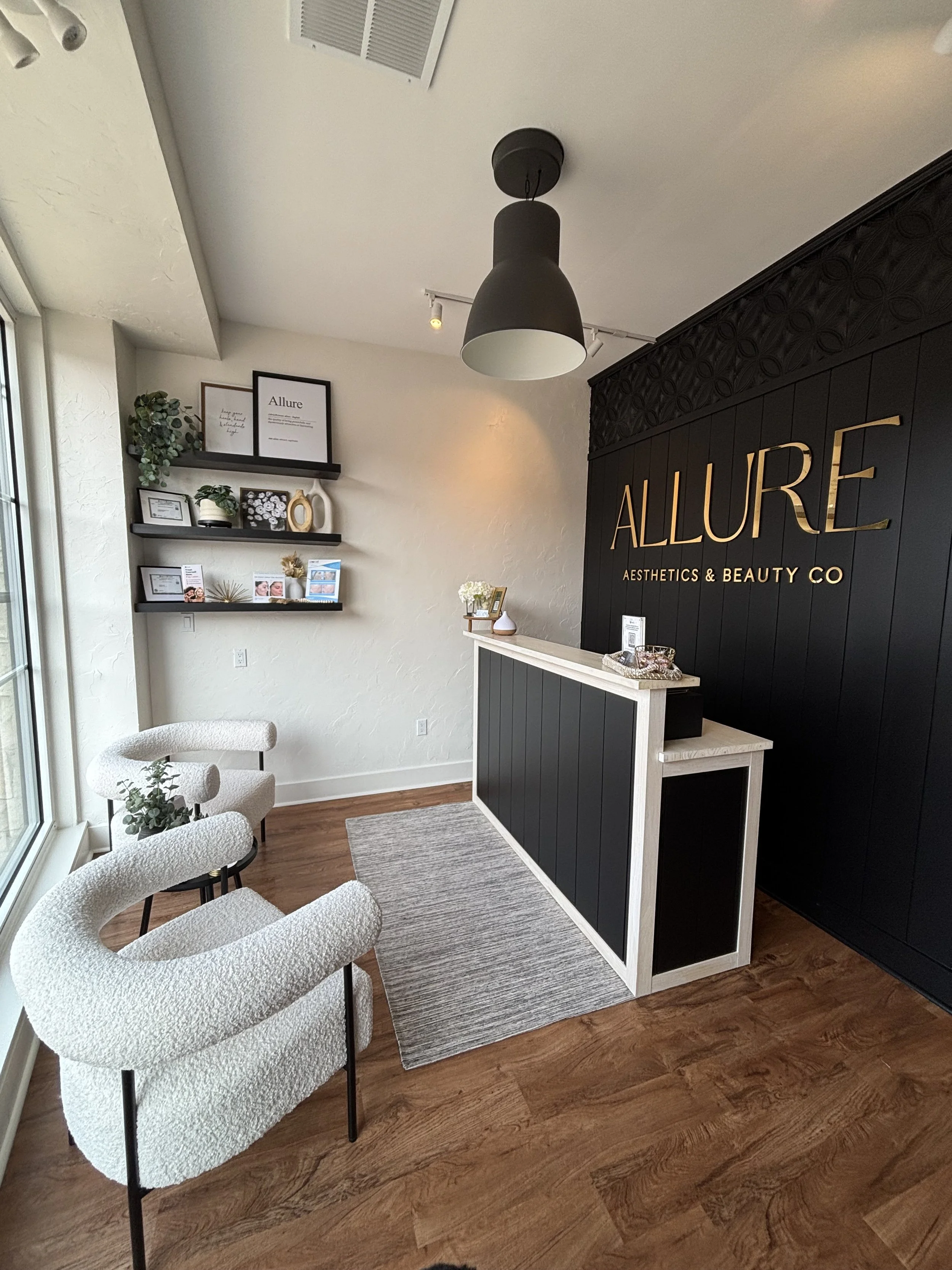 Interior of Allure Aesthetics & Beauty Co. reception area with black and white decor, chairs near window, a reception desk, wall shelves with picture frames and plants.