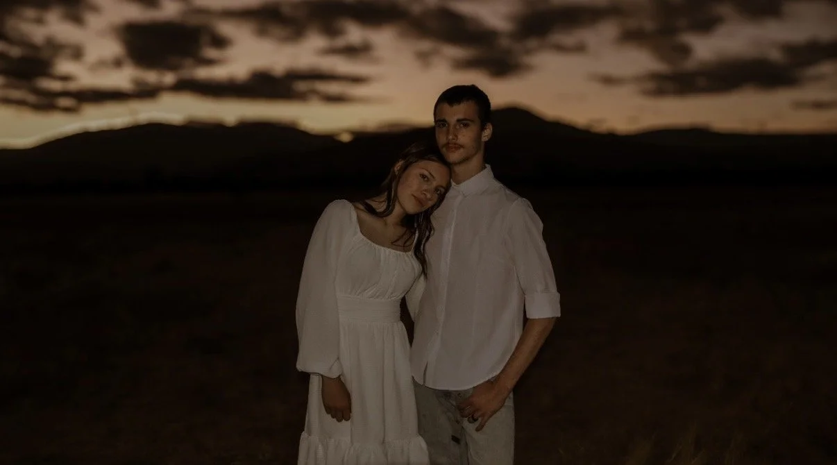 A young couple standing close together outdoors at sunset, with mountains in the background. The woman is leaning her head on the man's shoulder, and they are both dressed in white.