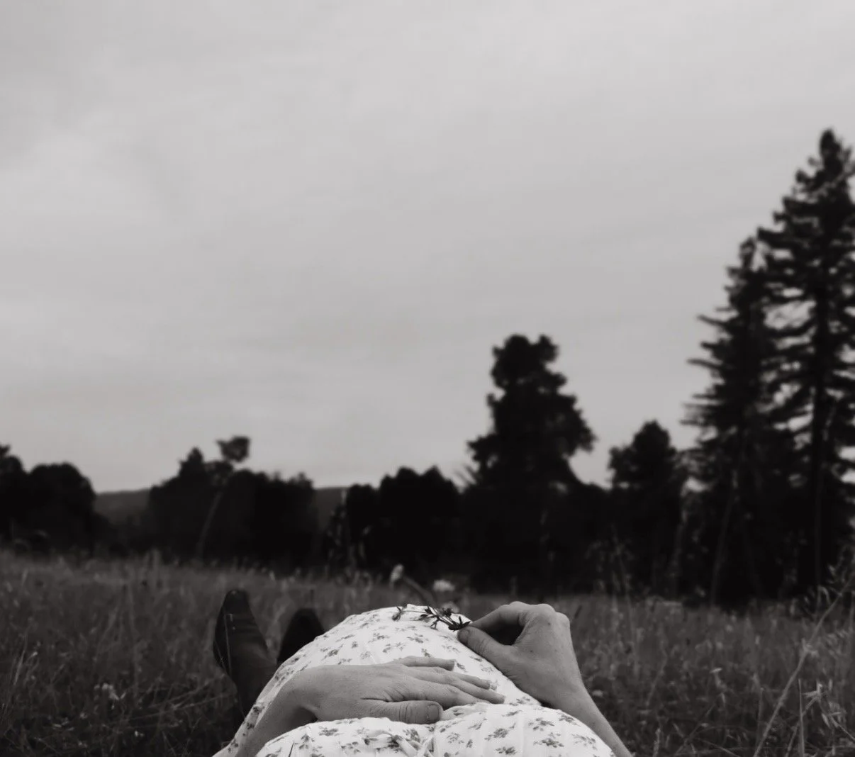 Person lying down in a grassy field holding a sprig of plant, with trees and cloudy sky in the background, in black and white.
