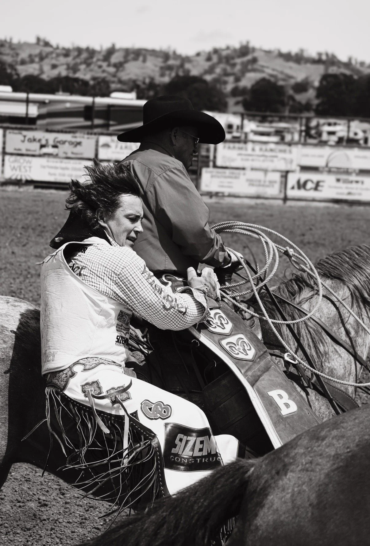 A woman and a man riding horses, with the woman's hair blowing in the wind, at an outdoor rodeo or horse riding event.