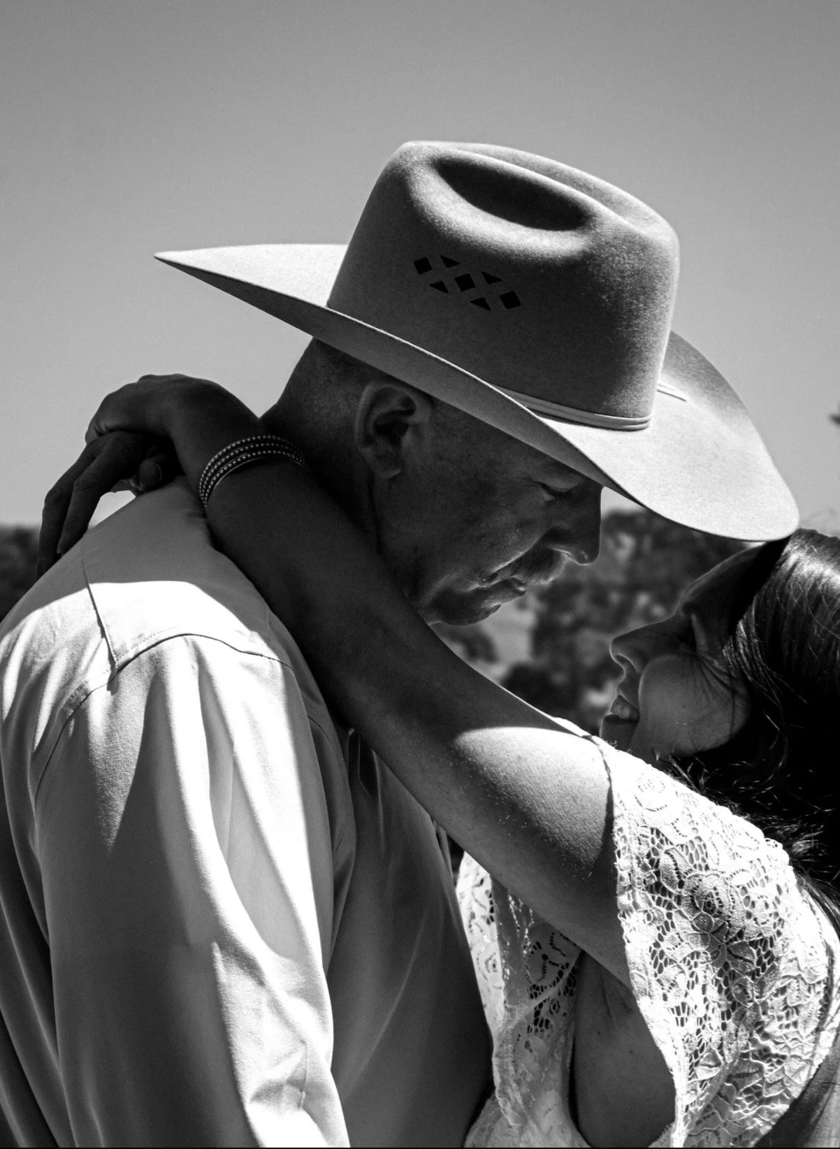 A black and white photo of a couple embracing, with the woman looking into the man's face, wearing a lace top and the man wearing a hat and a light-colored shirt.