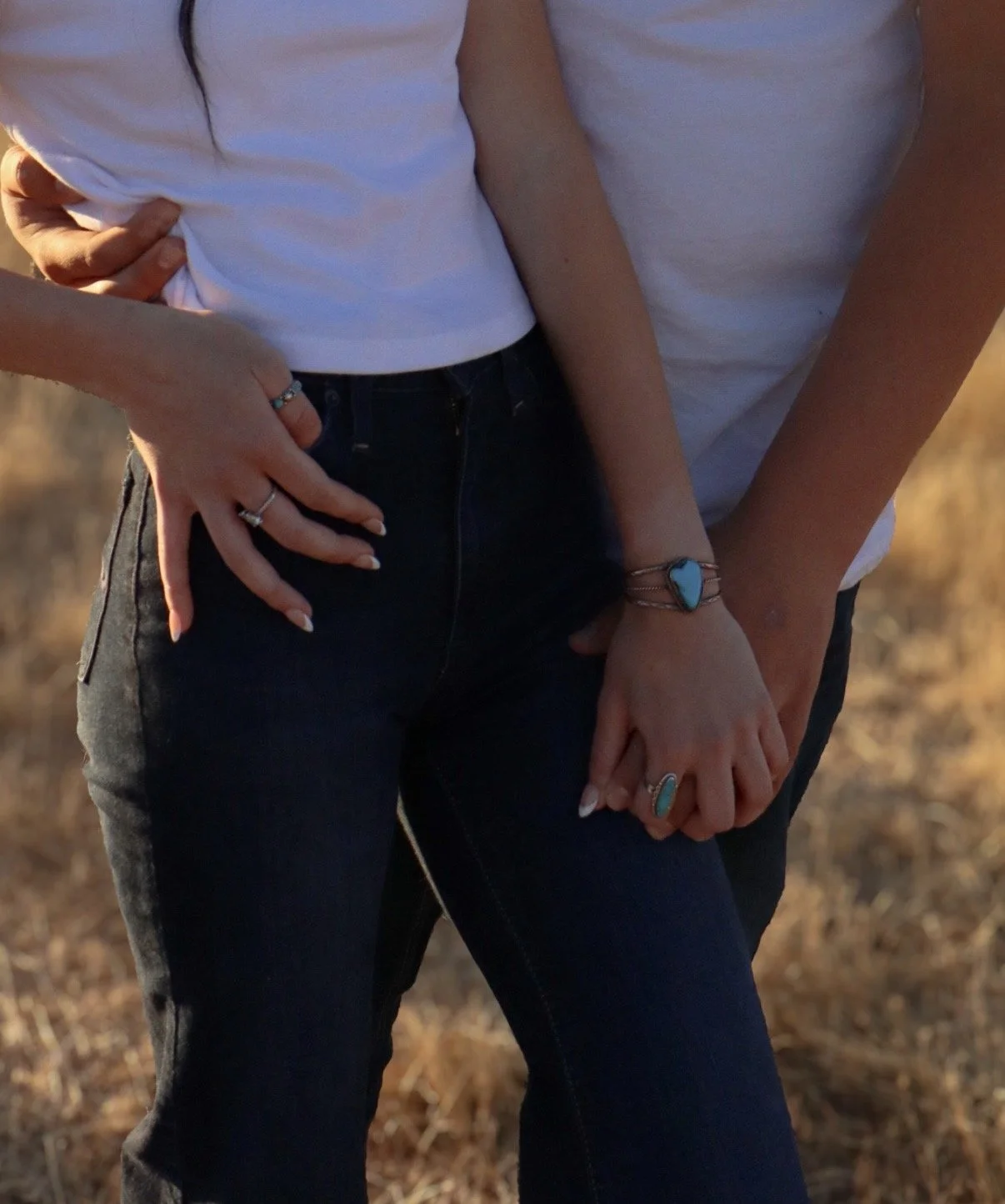 Close-up of two women holding hands, wearing white t-shirts and dark jeans, with jewelry including rings, bracelets, and rings with turquoise stones, outdoors on a dry, grassy field.