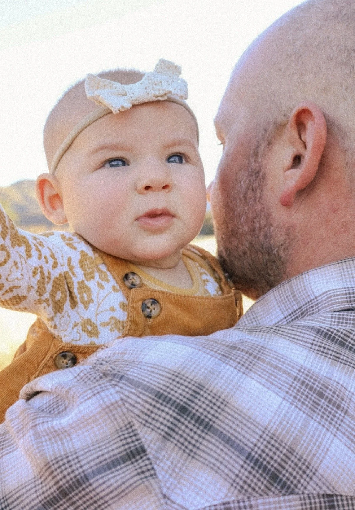 A close-up of a man holding a young child on his shoulder outdoors, with the man facing away and the child's face visible, looking slightly to the side. The child has blue eyes, light skin, and is wearing a cream-colored headband with a bow and a mustard-colored outfit with a patterned shirt.
