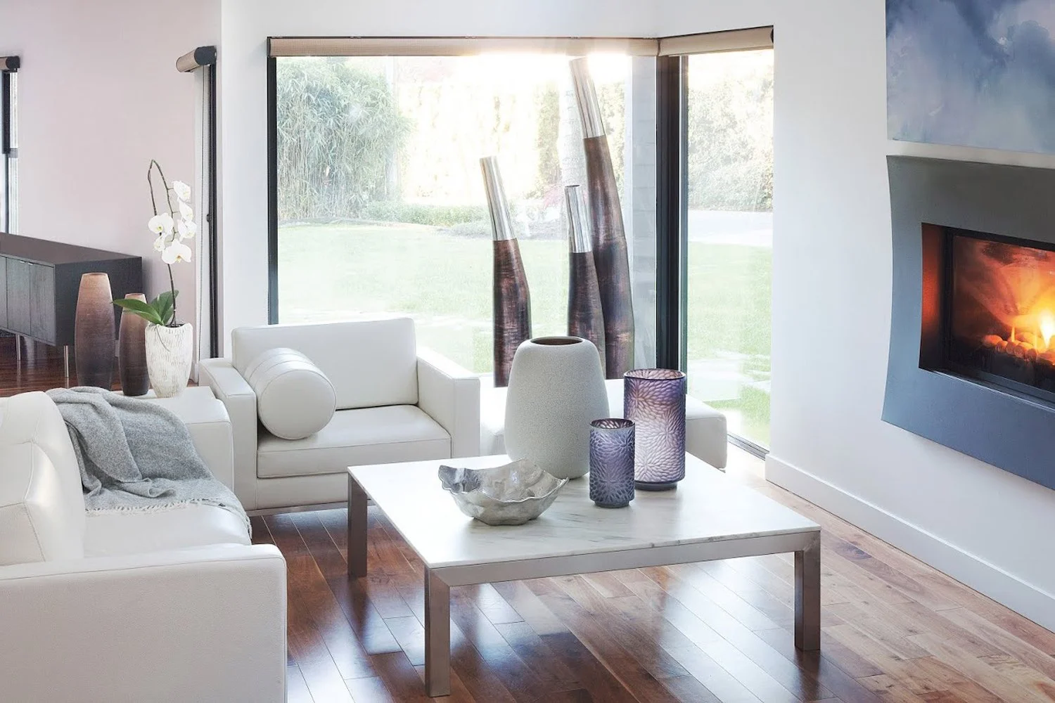 Modern living room with white sofas, marble coffee table, decorative vases, and a fireplace. Large windows offer garden views and natural light.