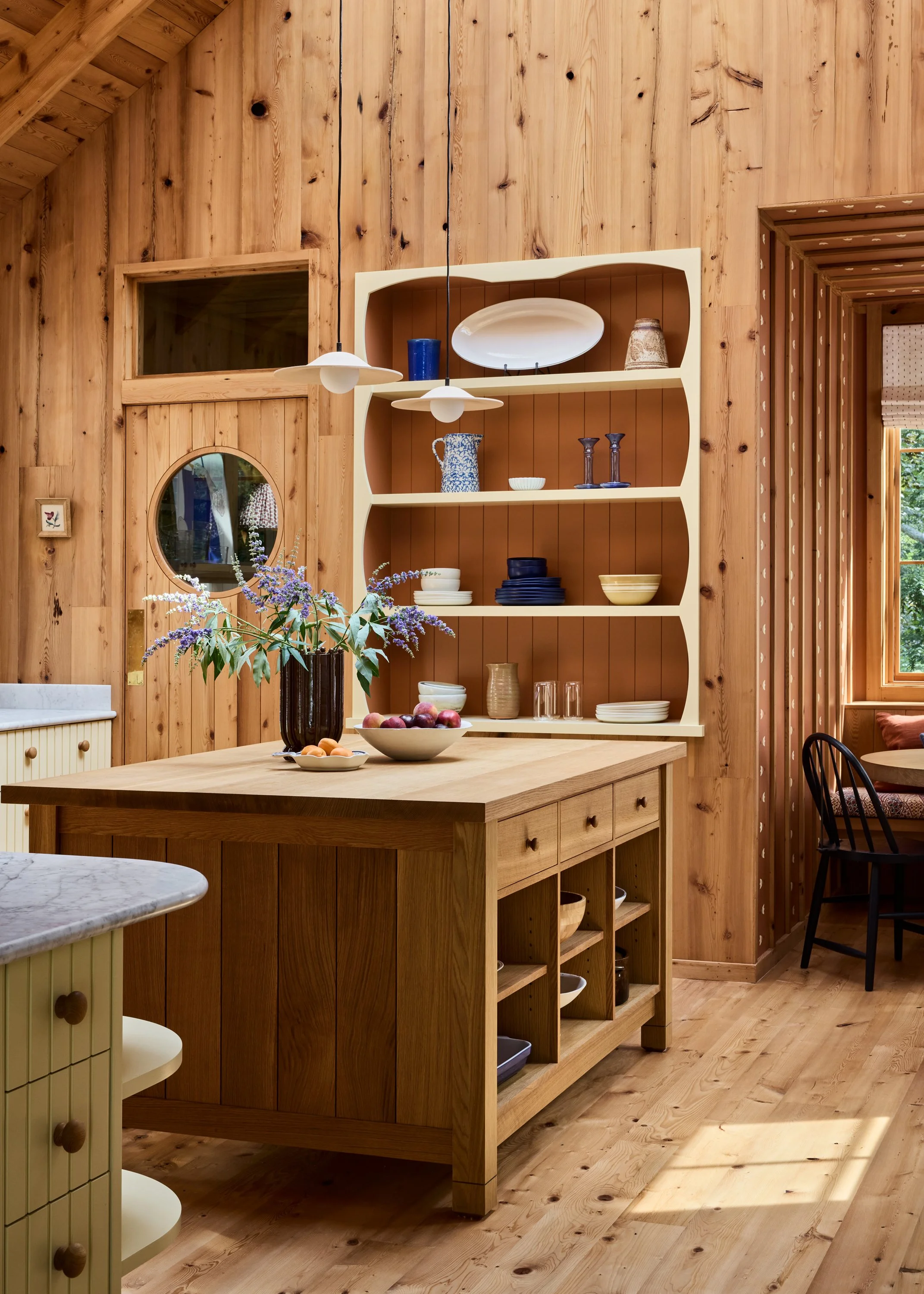 Wood-paneled kitchen with a central island, open shelving with dishes, and a small adjoining table with a black chair, decorated with a flower arrangement.
