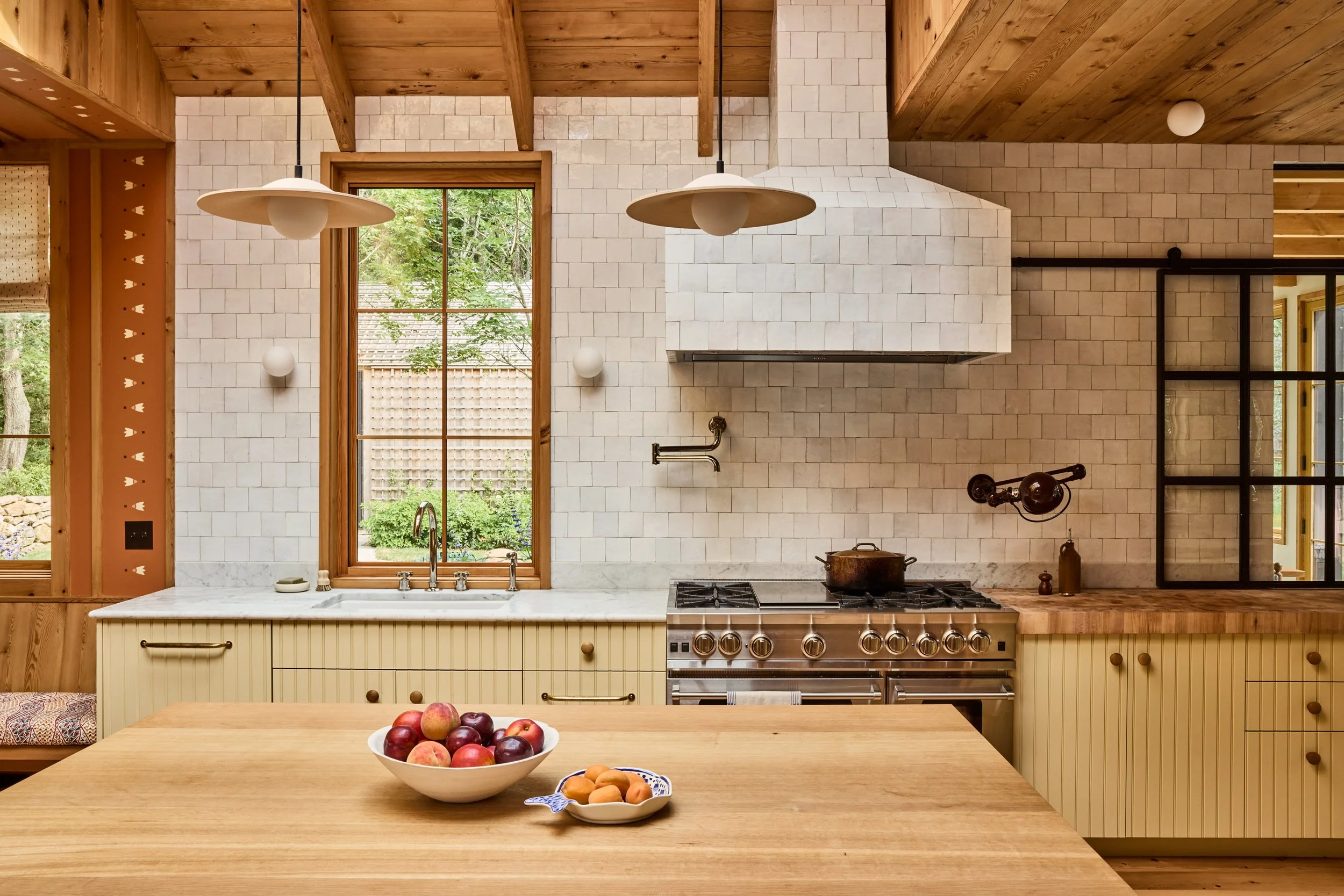 A rustic kitchen with cream-colored cabinets, a wooden countertop, and a central wooden table topped with a bowl of apples and a small dish of apricots. The kitchen features a large window, a modern stove, and hanging pendant lights.