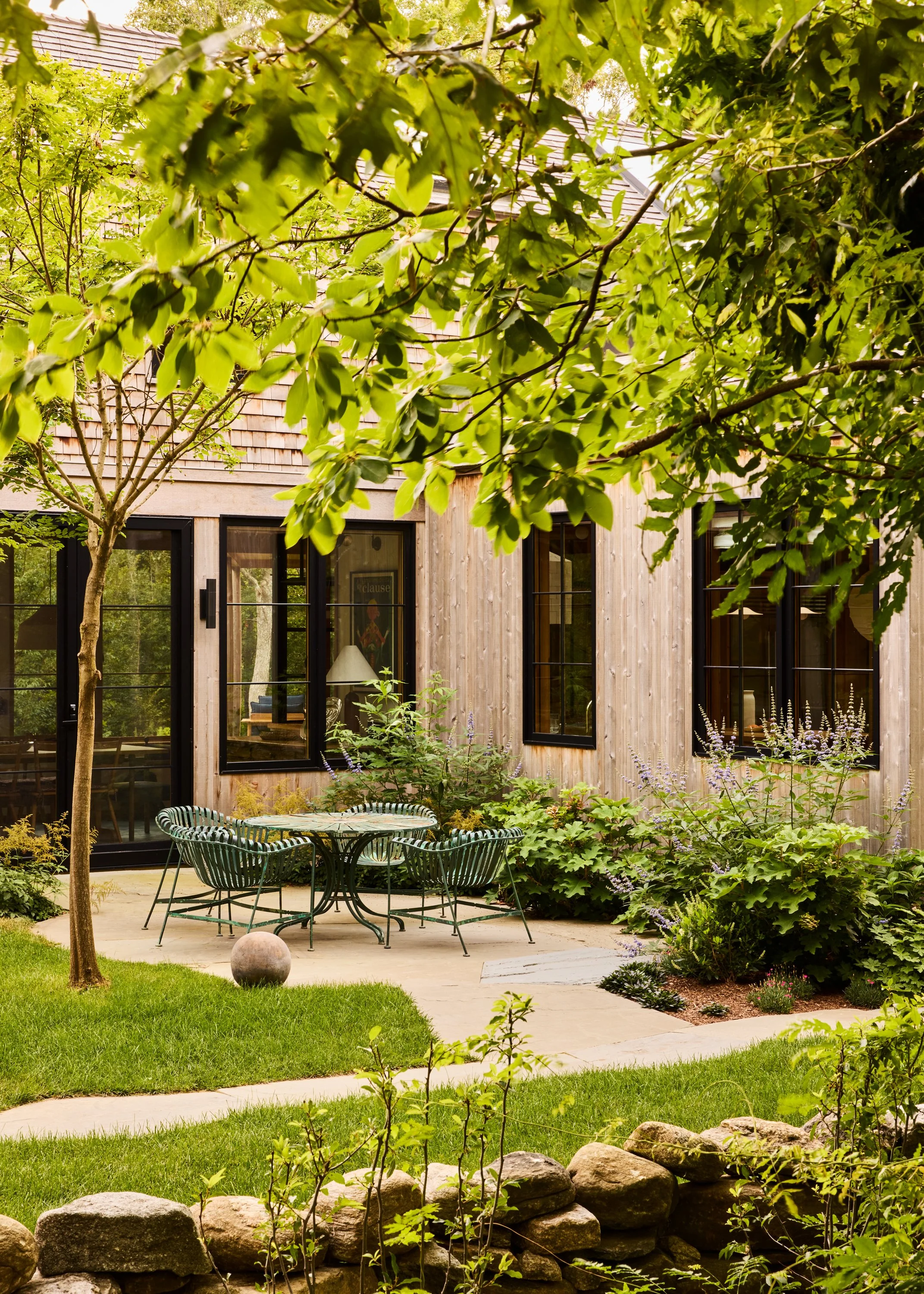 A backyard patio with a round metal table and six metal chairs surrounded by lush green plants and trees, with a wooden house with large black-framed windows in the background.